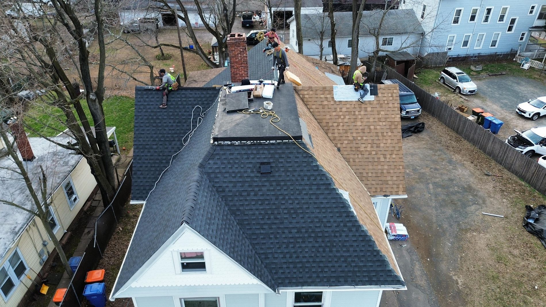 Rooftop work in progress on a house, with black shingles, tan plywood, and workers visible on the roof.