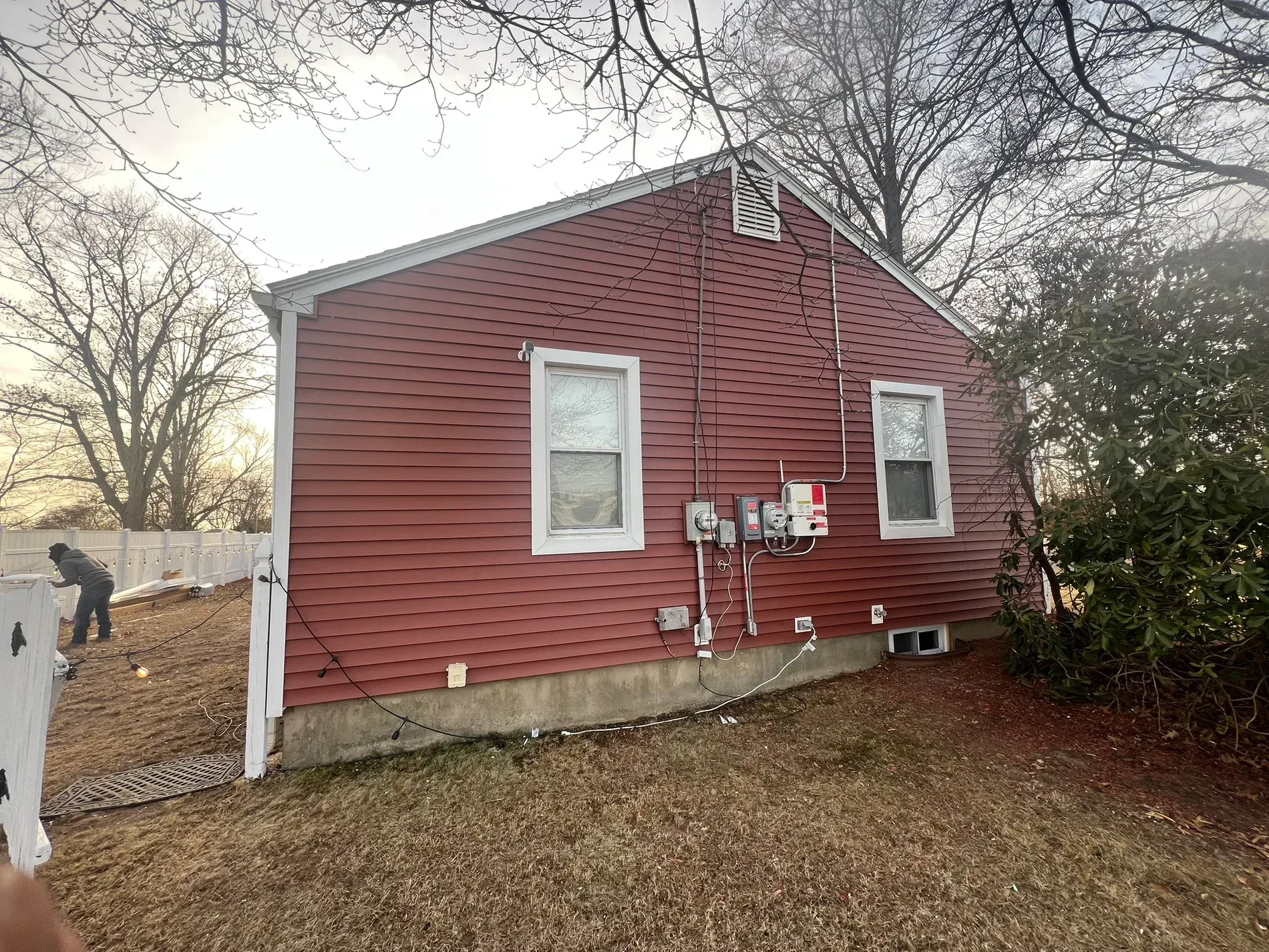 Small red house exterior with white trim, two windows, utility meters, and a fenced yard in late fall.