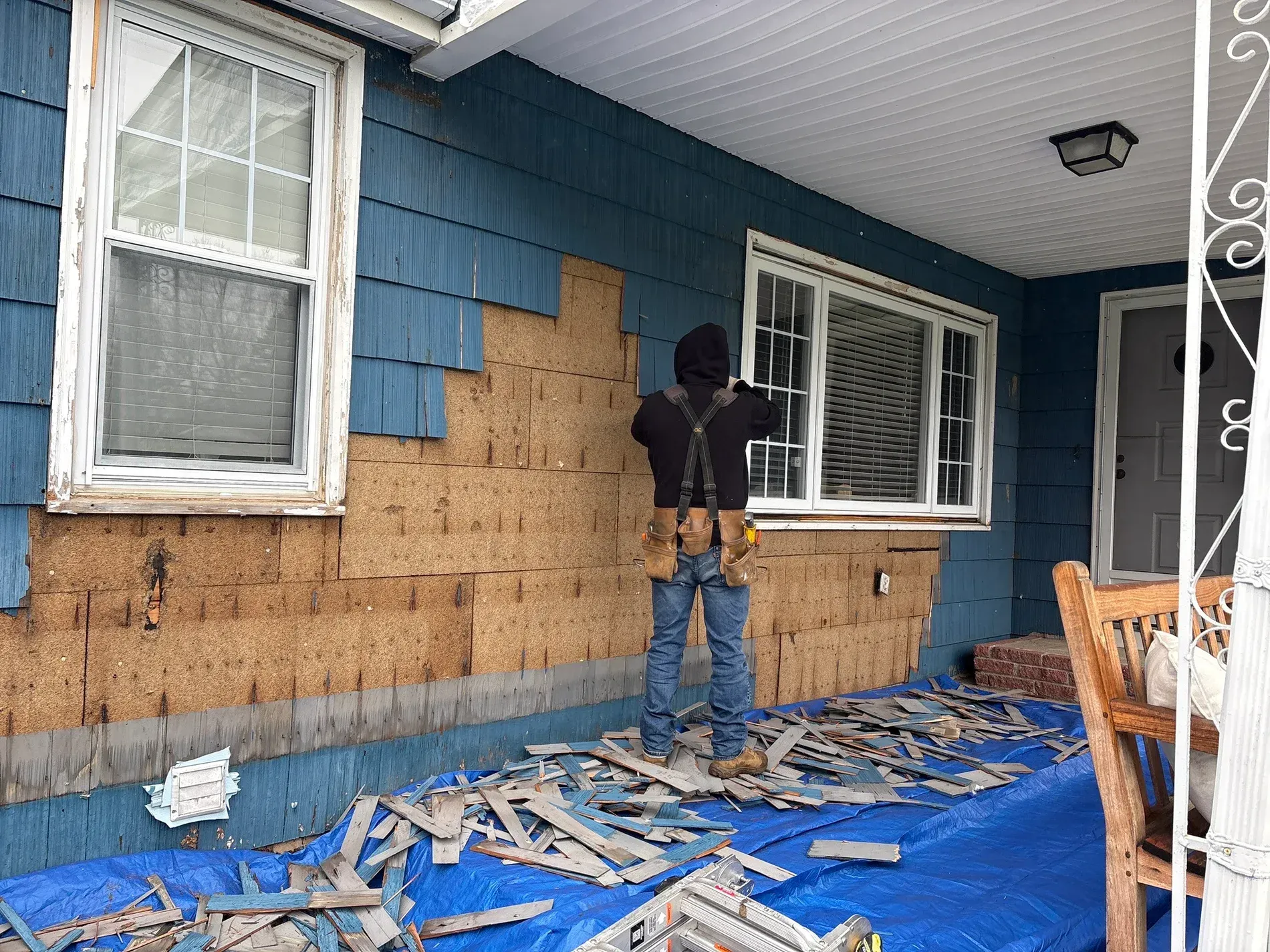Person repairs damaged siding on a blue house porch, with tools and debris on a tarp.