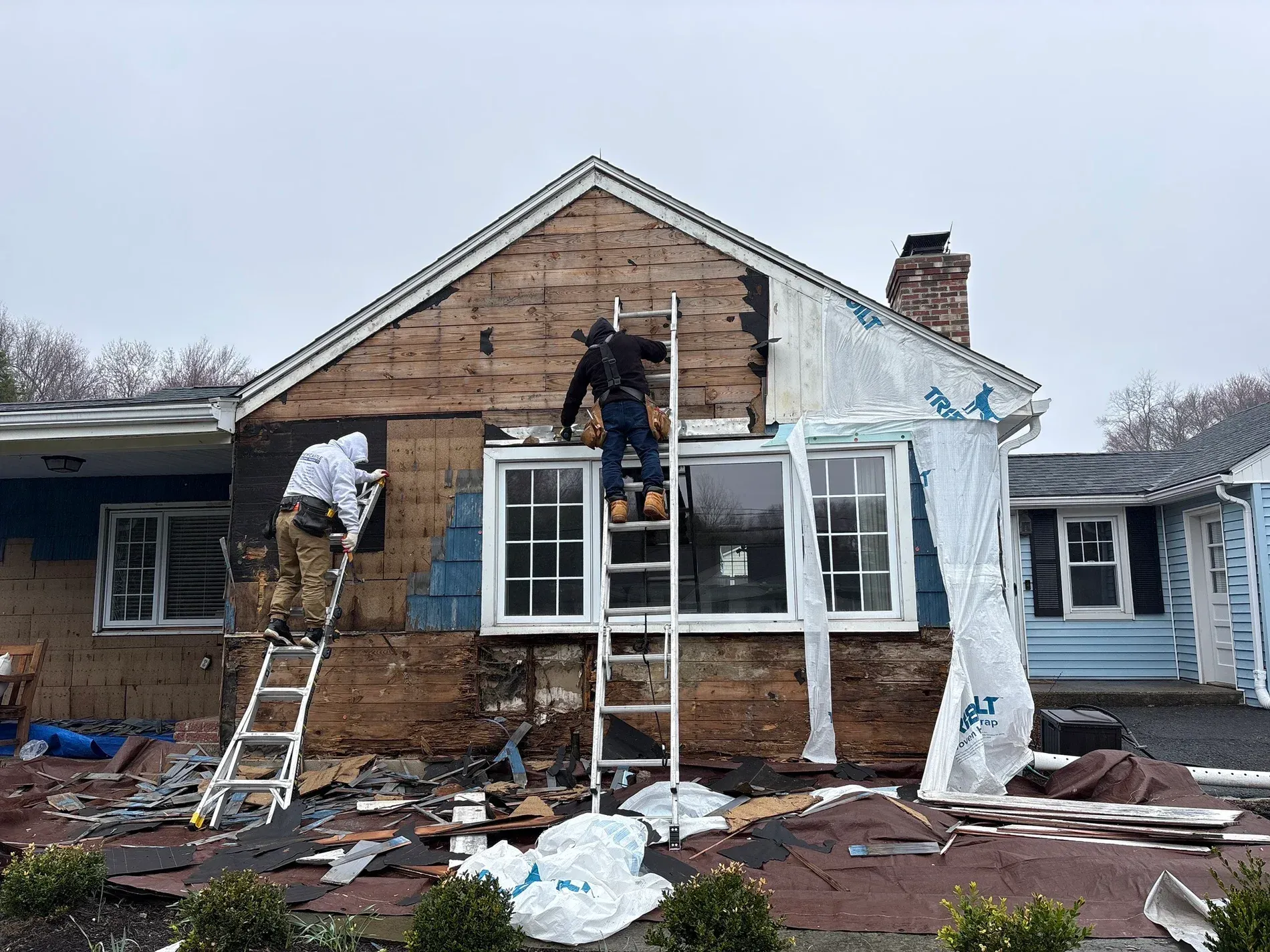 Workers repairing storm-damaged house roof with ladders, exposed siding, and debris on the ground.