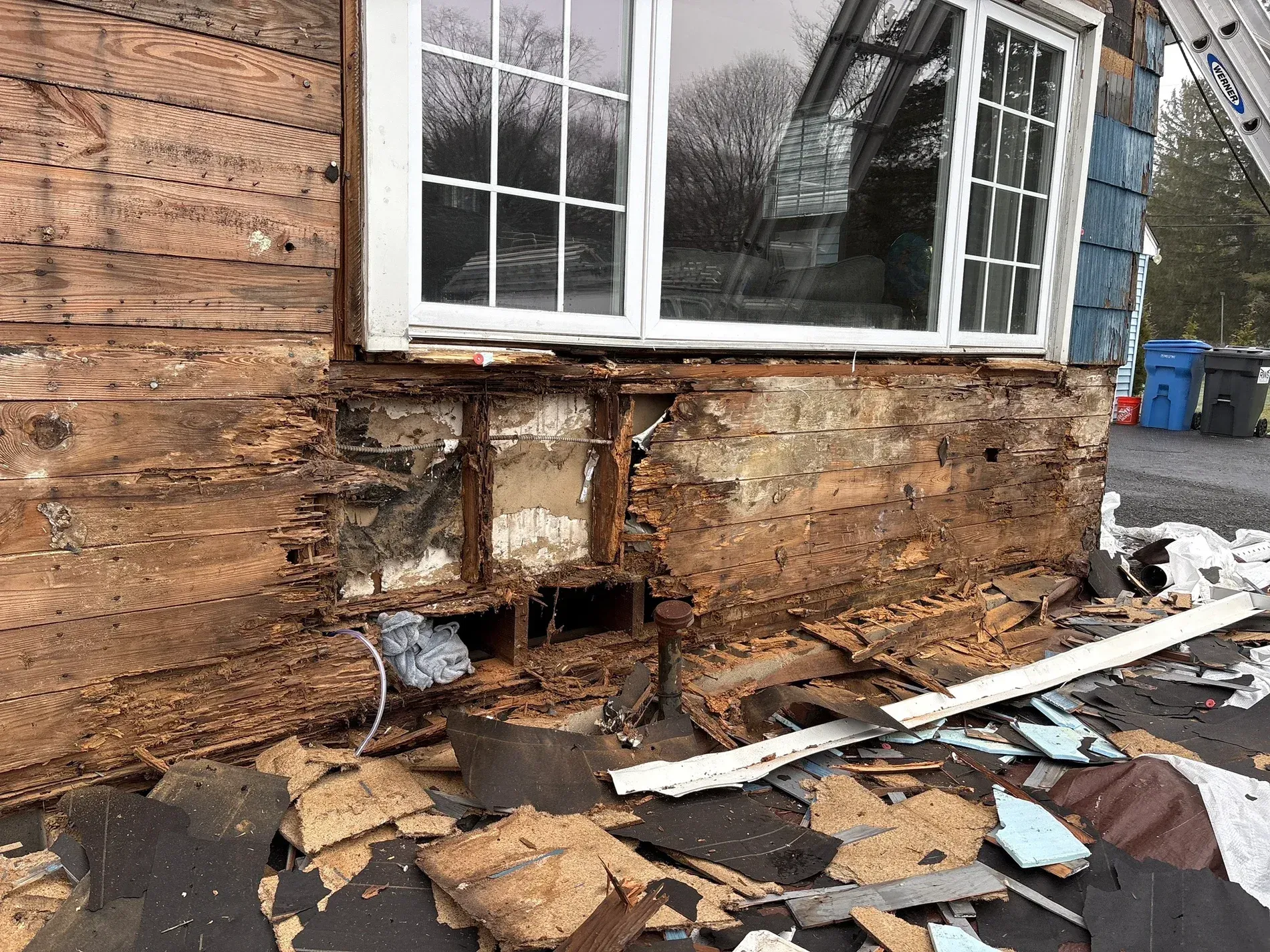 Damaged exterior wall of a house with exposed wood, broken siding, and debris beneath a window
