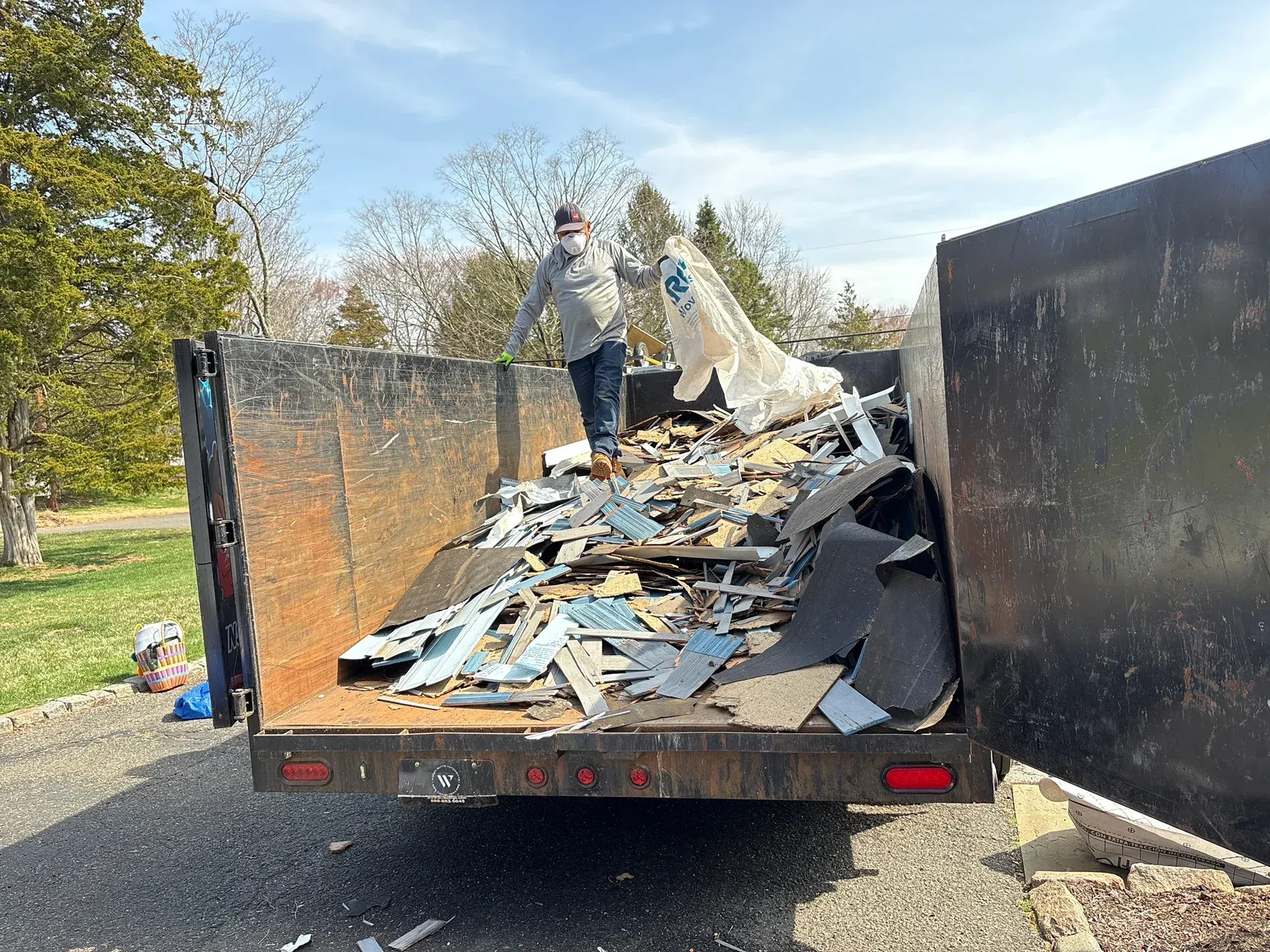 Two workers loading debris into an open trailer in a yard, with a large pile of scrap inside.