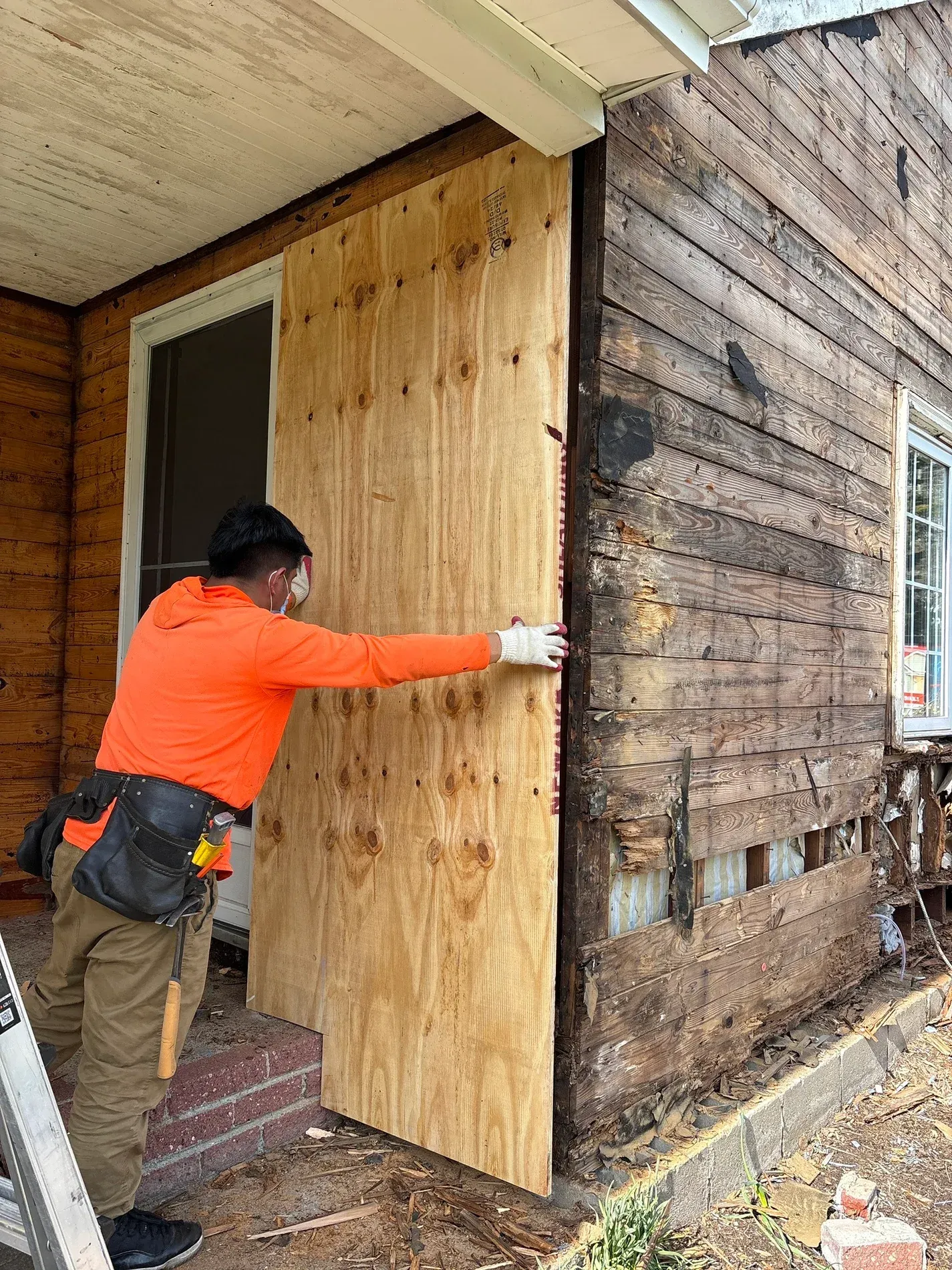 Worker in orange shirt installing plywood over a damaged exterior wall during home repair.