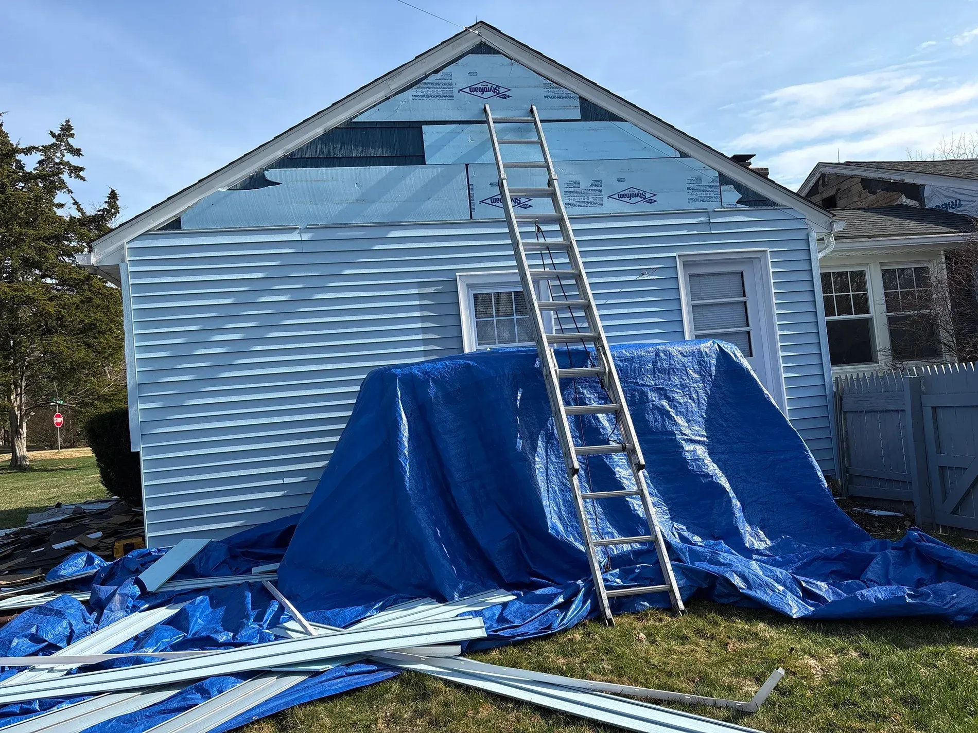 Blue house with a ladder on the roof and blue tarp covering the front yard work area