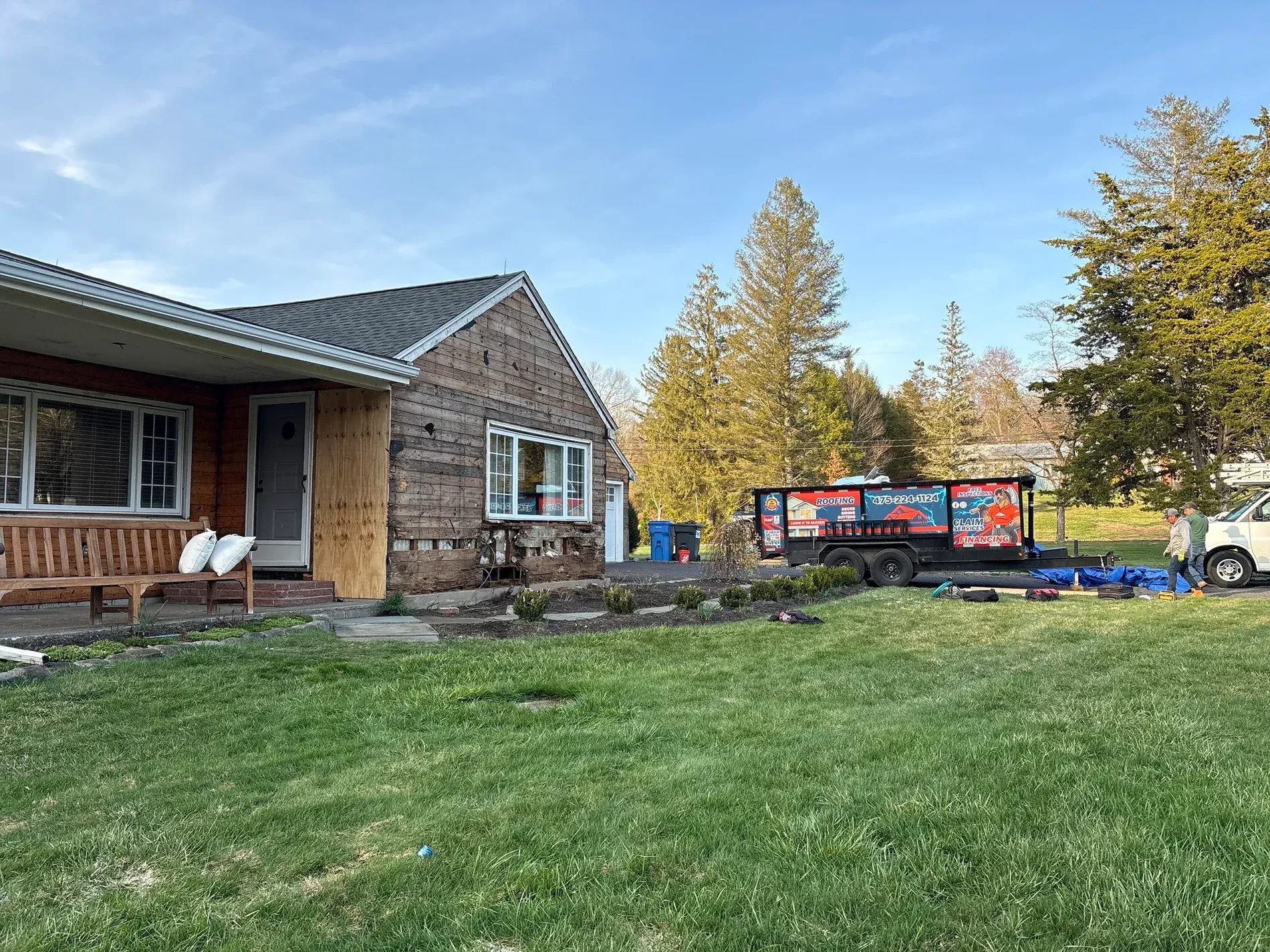 Wooden house with porch and lawn, autumn trees, and a parked blue trailer in the yard.