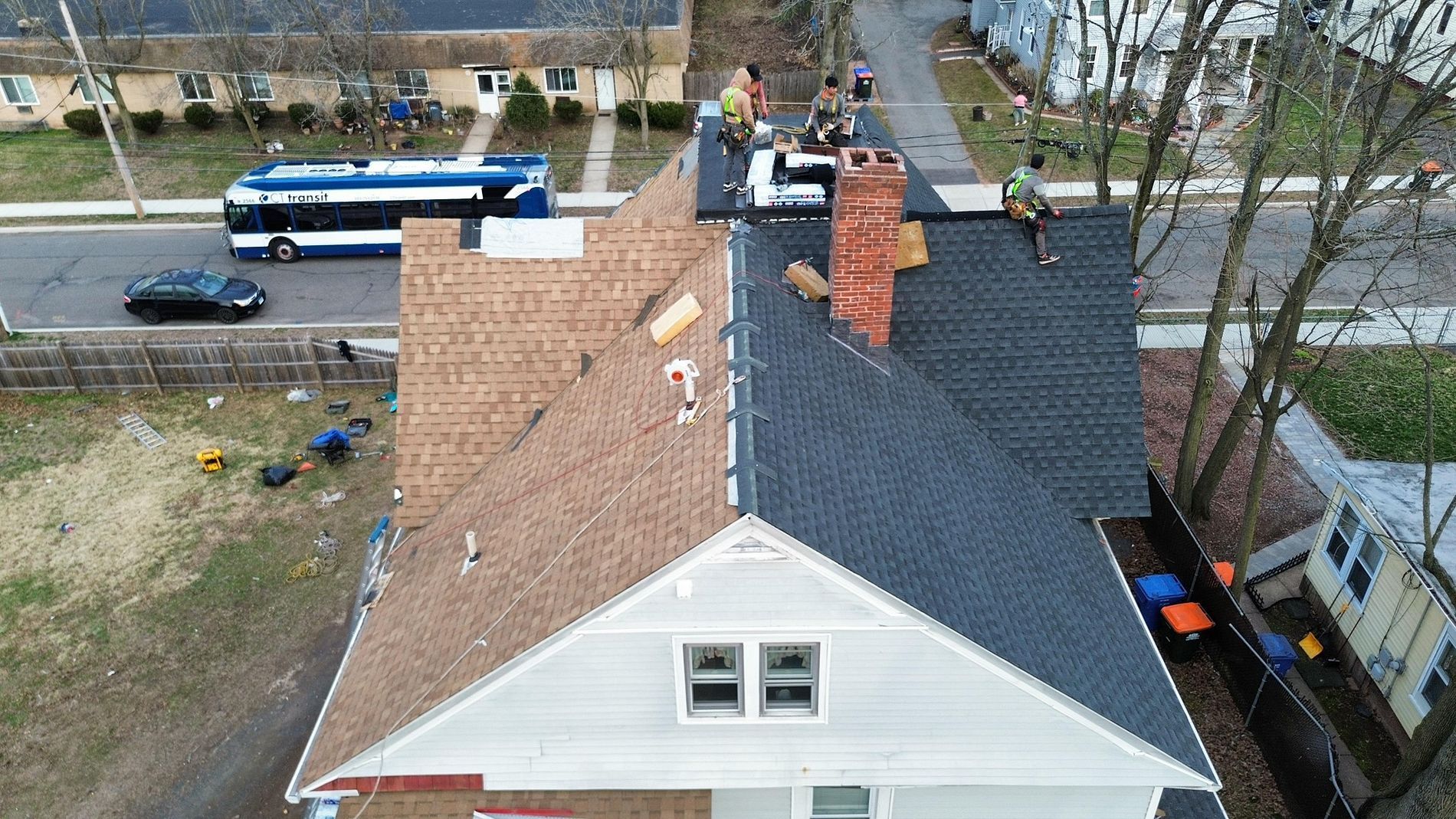 Aerial view of a house roof with shingles and two chimneys beside a street and parked bus.