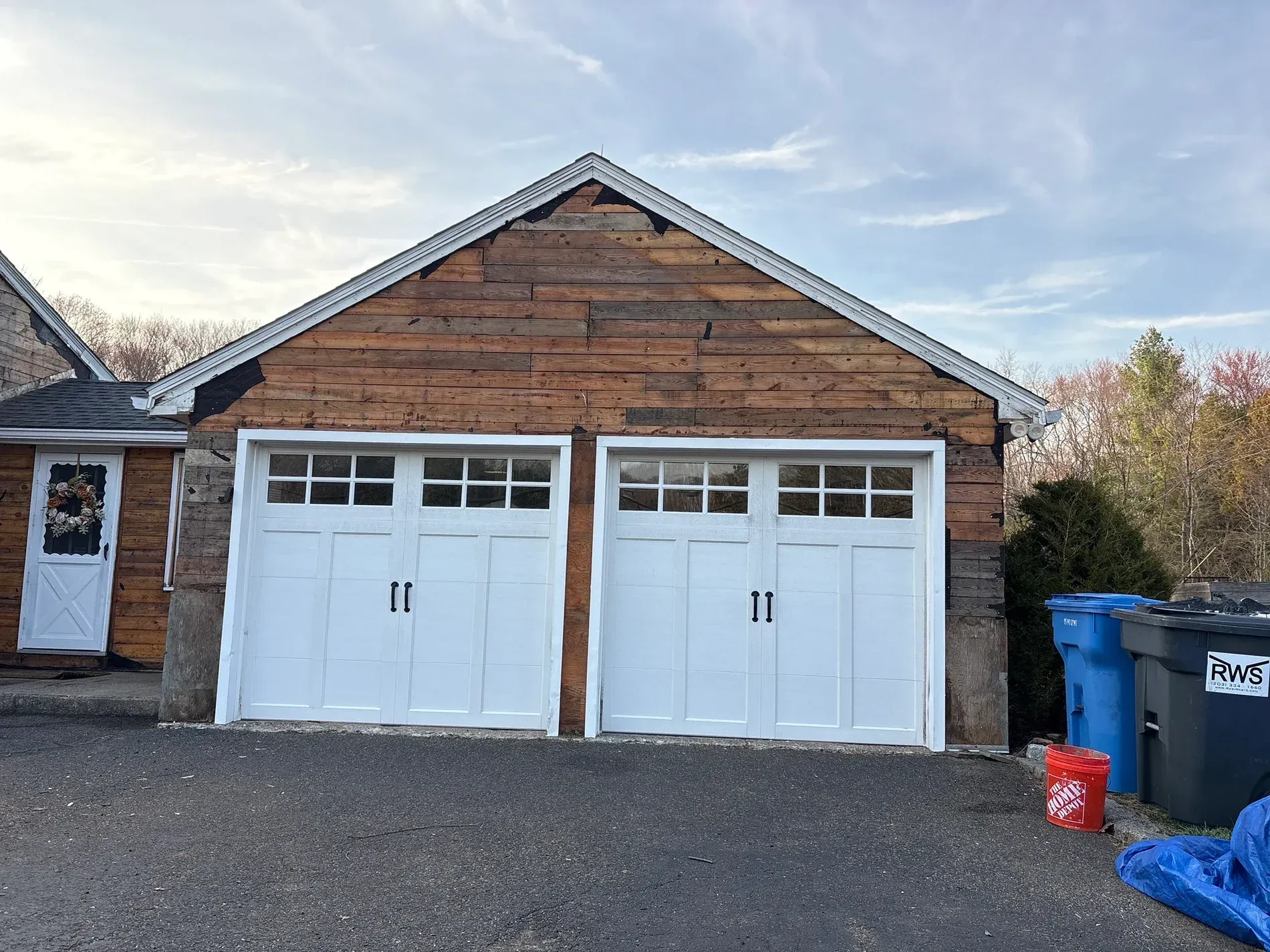 Brick garage with two white doors, gravel driveway, and trash bins on the right.