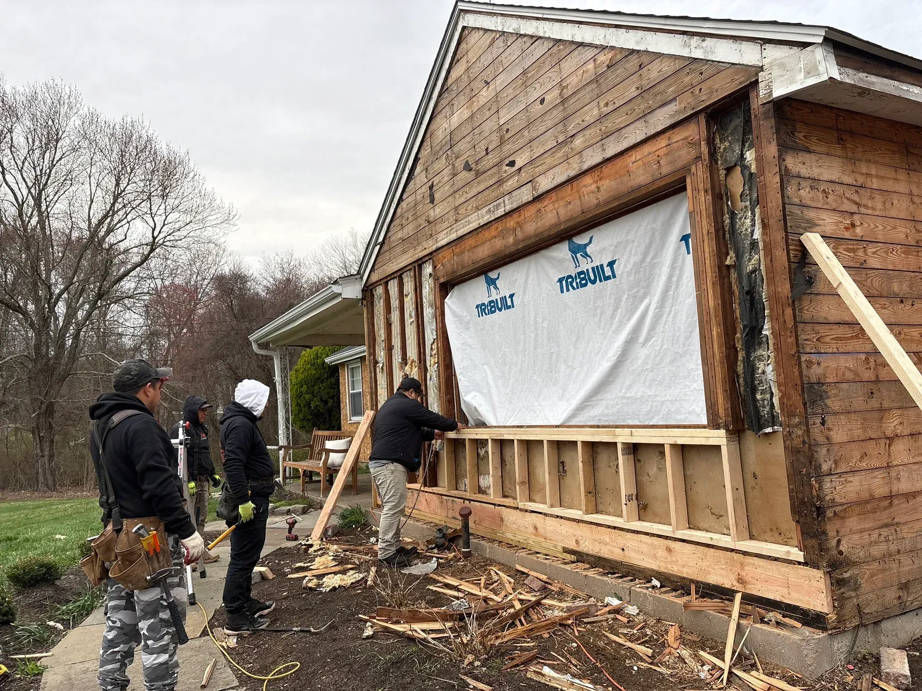 Workers framing and sheathing a house exterior during construction on a wet, wooded lot.