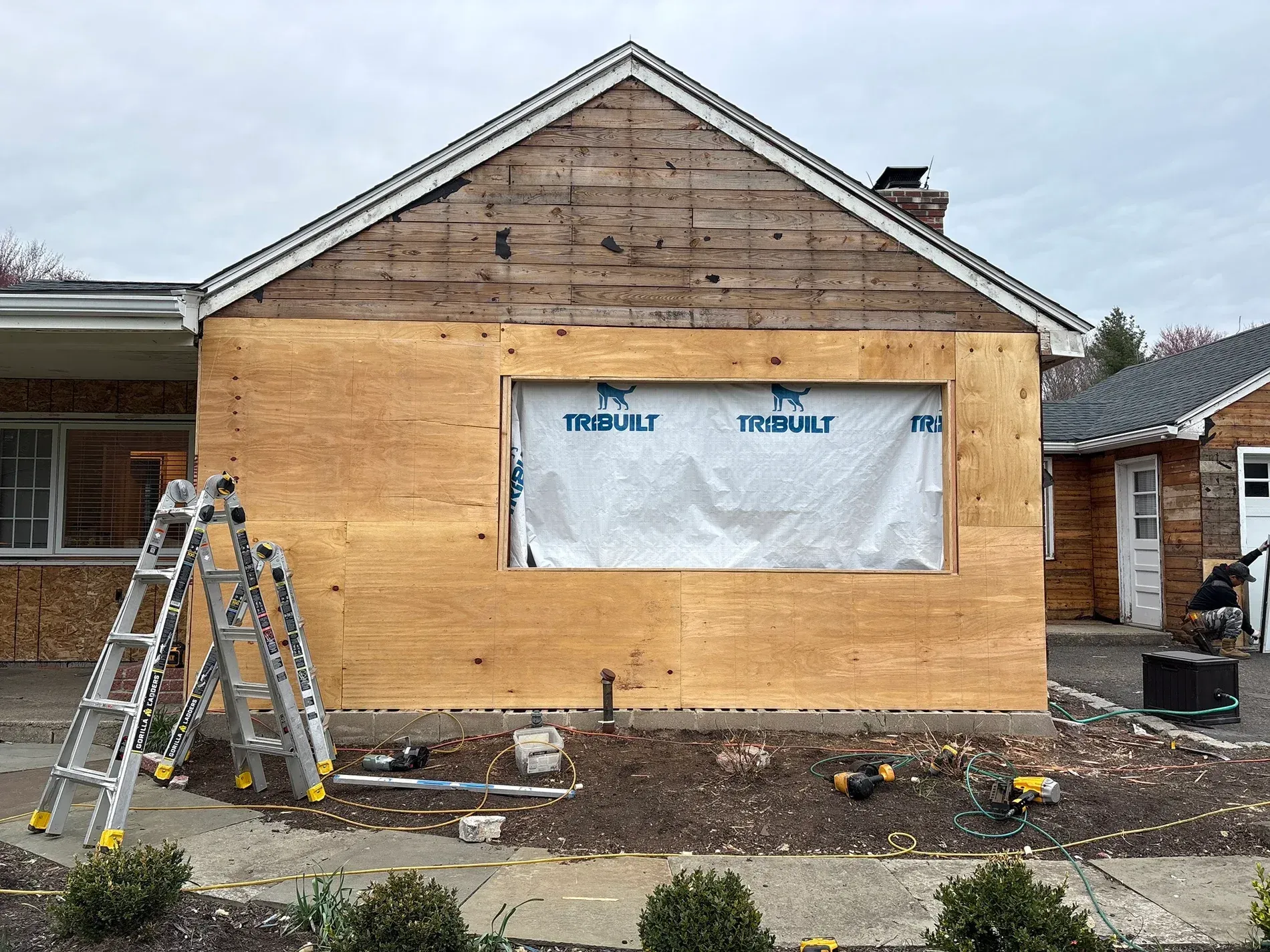 House exterior under renovation with plywood siding, covered window, and ladders in front