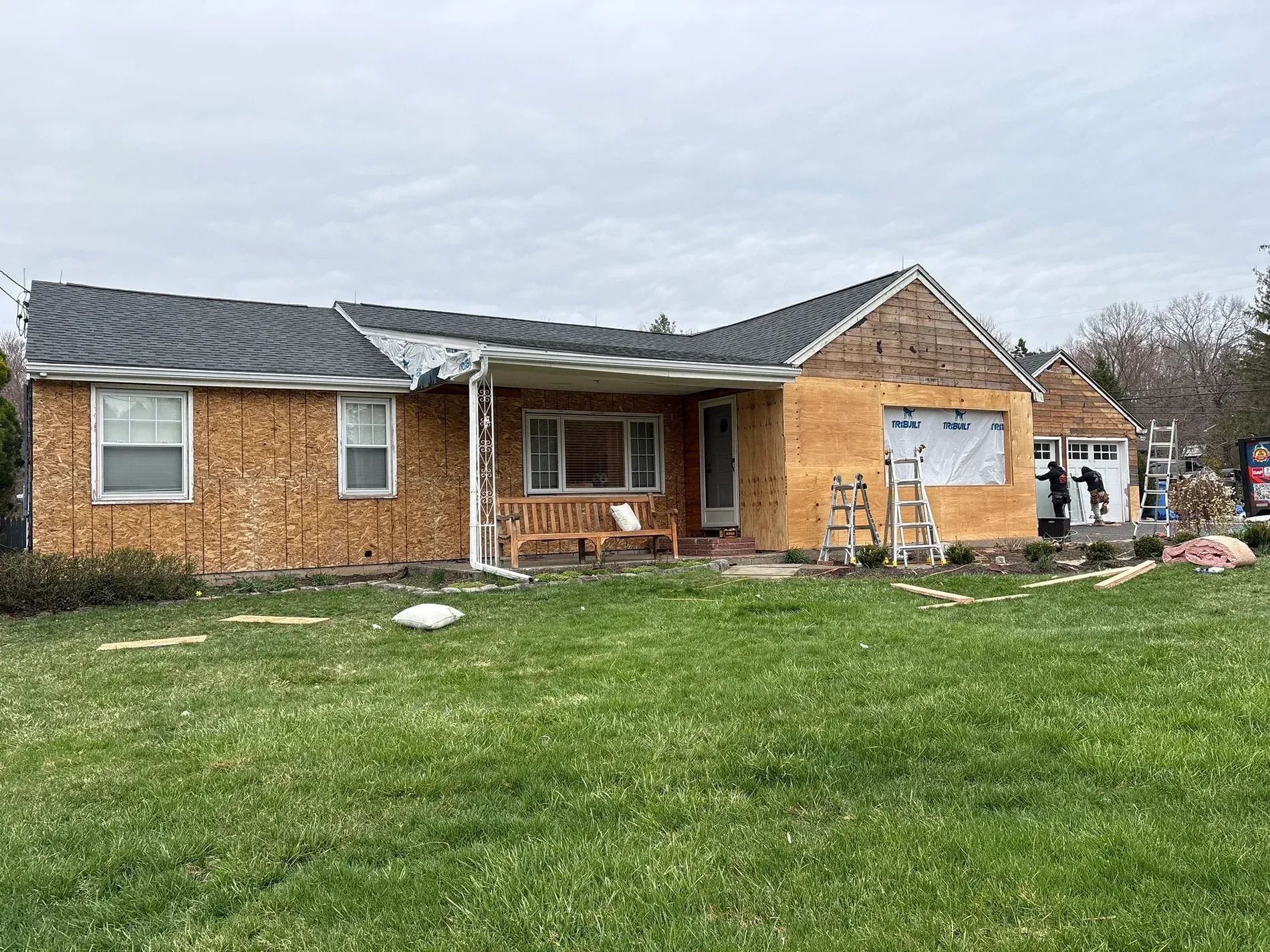 Damaged brick house with exposed wood siding, boarded window, and a debris-covered lawn
