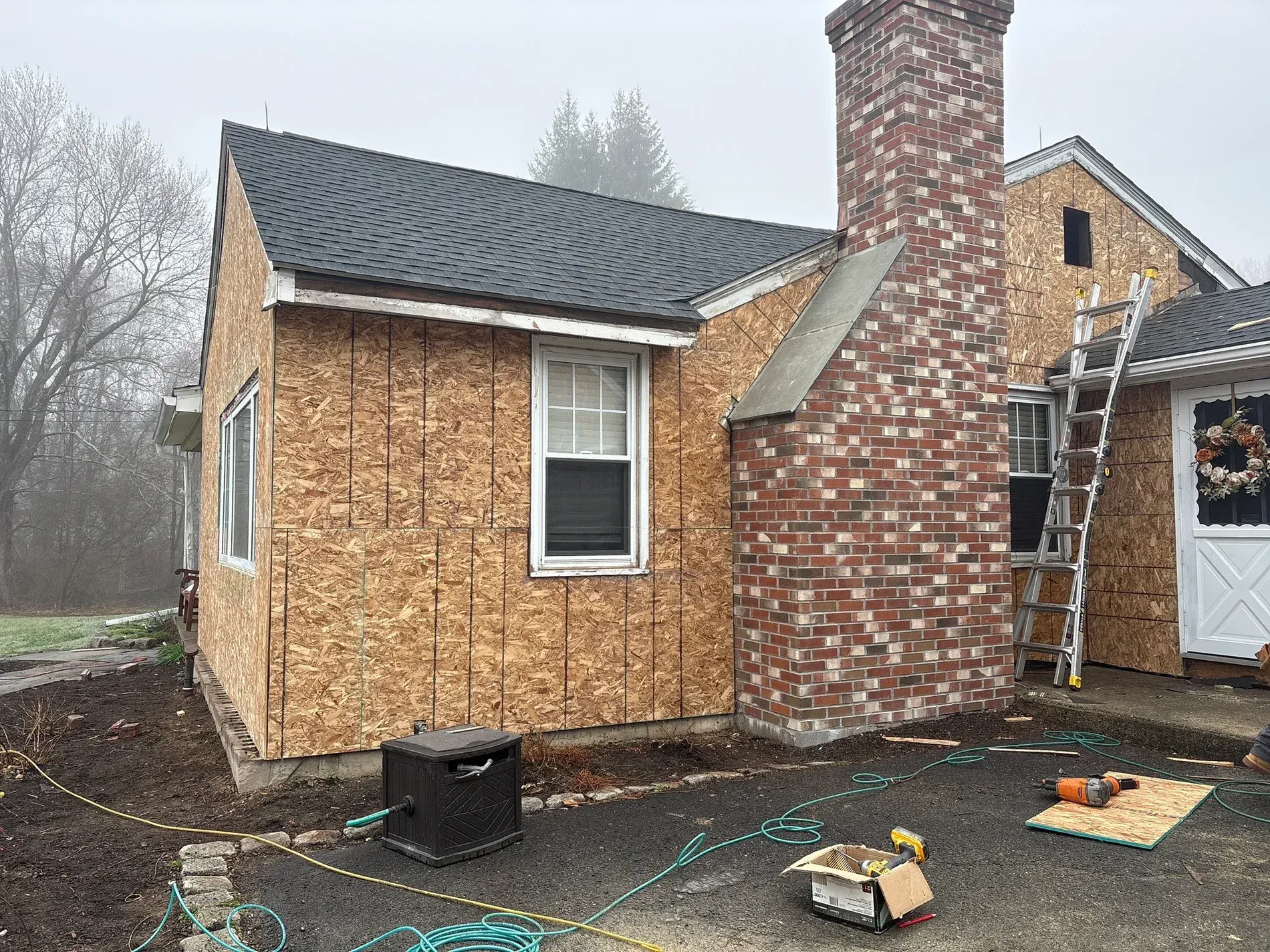Small house under construction with OSB siding, brick chimney, and roofing materials on the ground.
