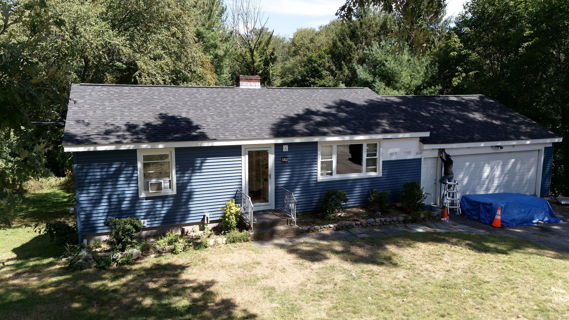 Blue single-story house with gray roof and a front porch, surrounded by trees and a grassy yard