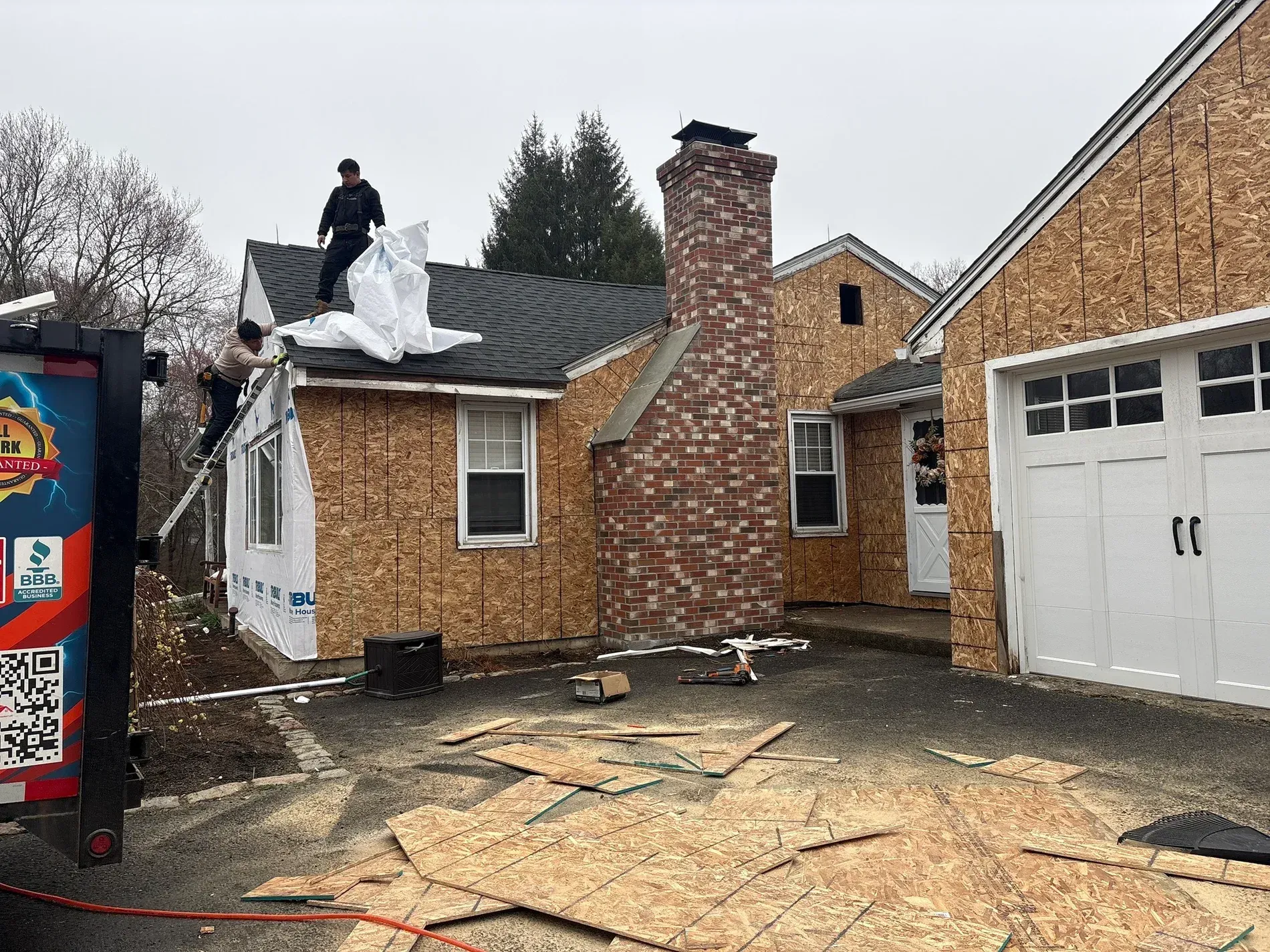 Worker on a roof with torn shingles beside a brick house and garage after storm damage.