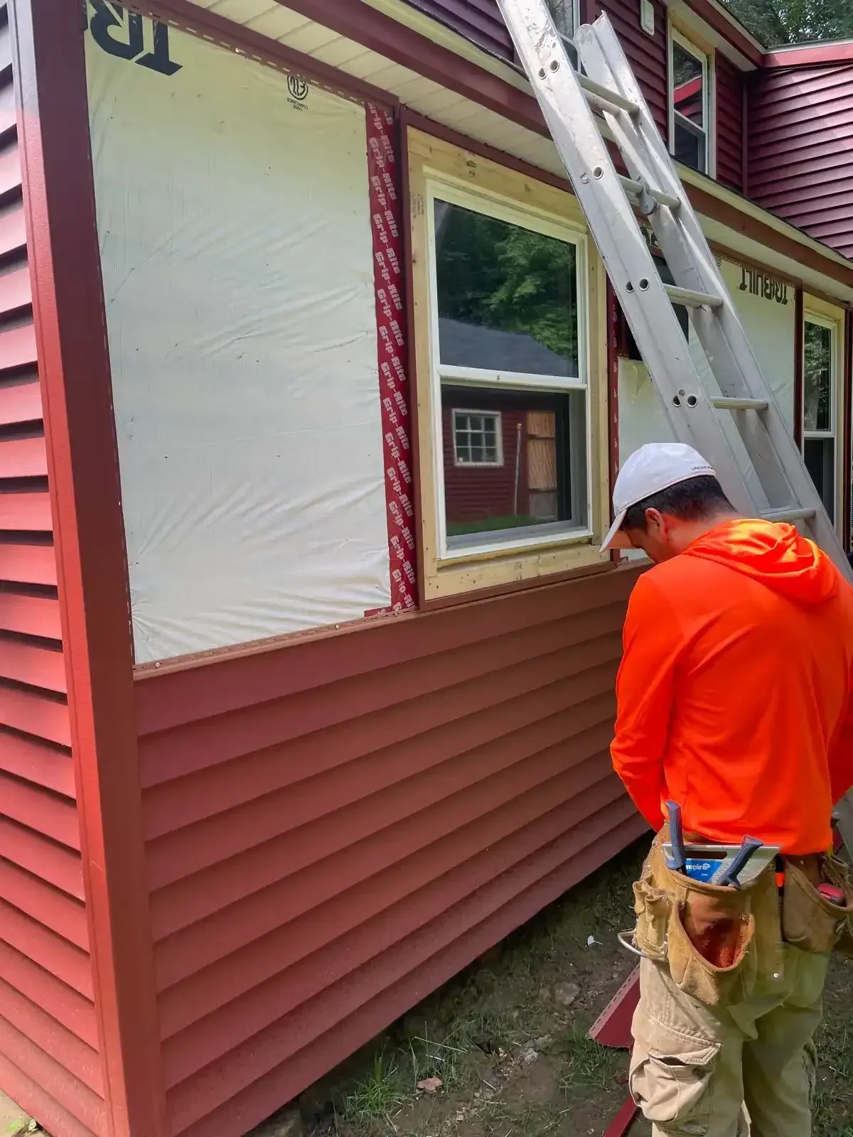 Worker in bright orange hoodie and white cap standing beside a house with ladders and covered windows.