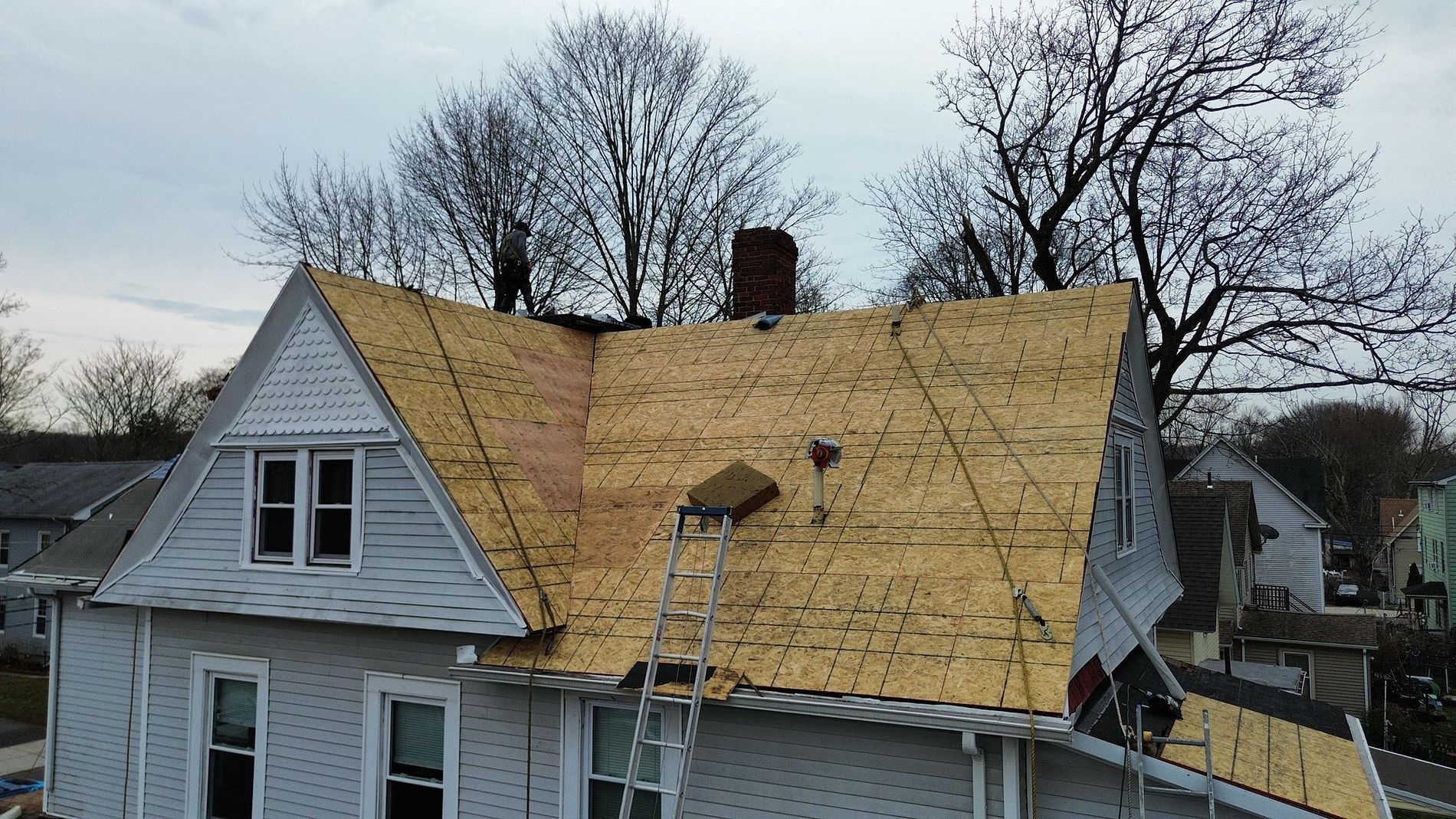 House roof under repair with workers on a steep plywood roof and surrounding trees in the background