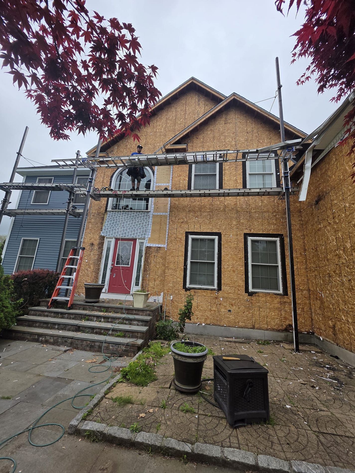Worker on scaffolding outside a tan house with red door and steps, surrounded by trees and construction materials