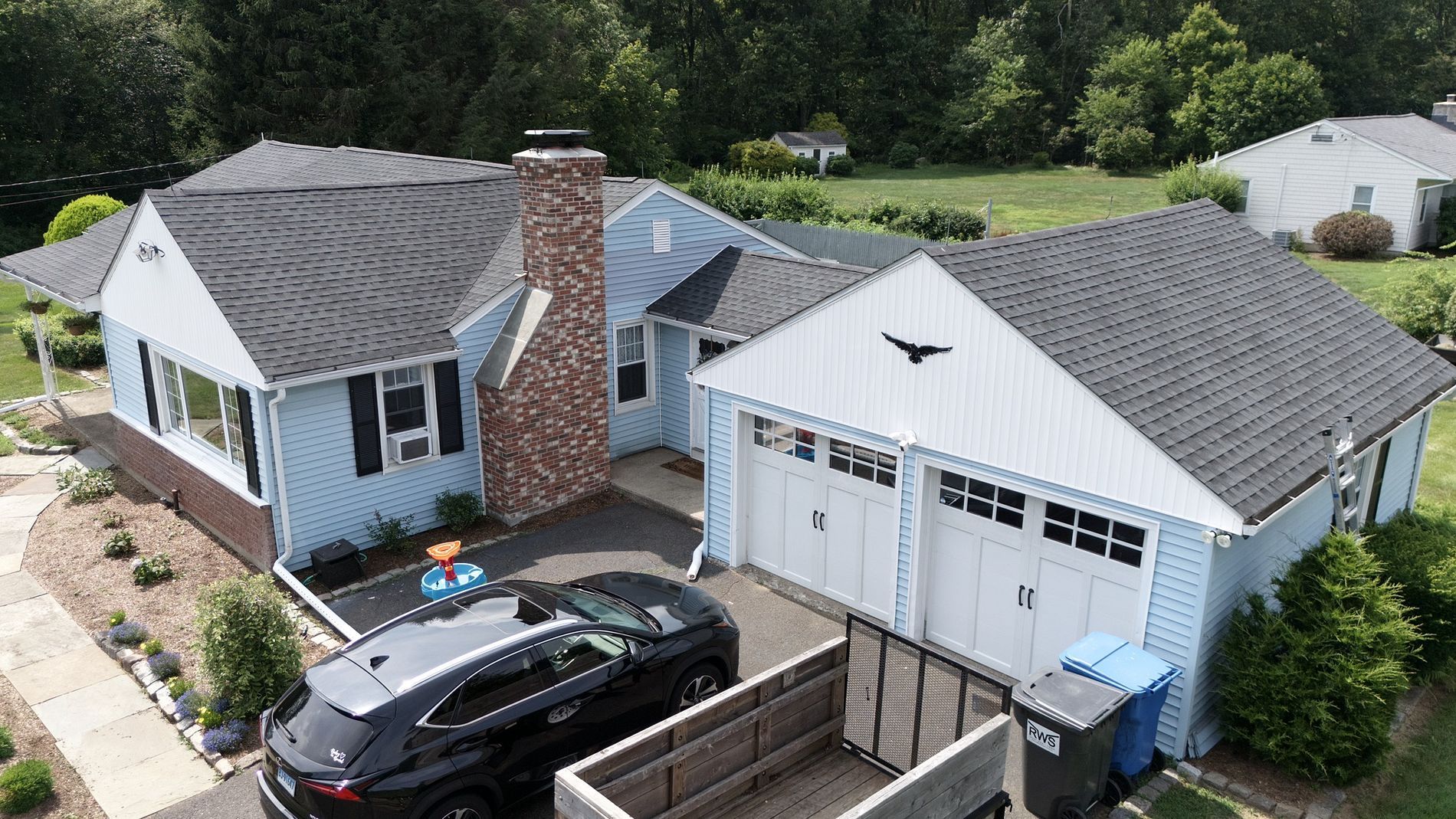 Aerial view of a white house and detached garage with a black car in the driveway and green trees nearby