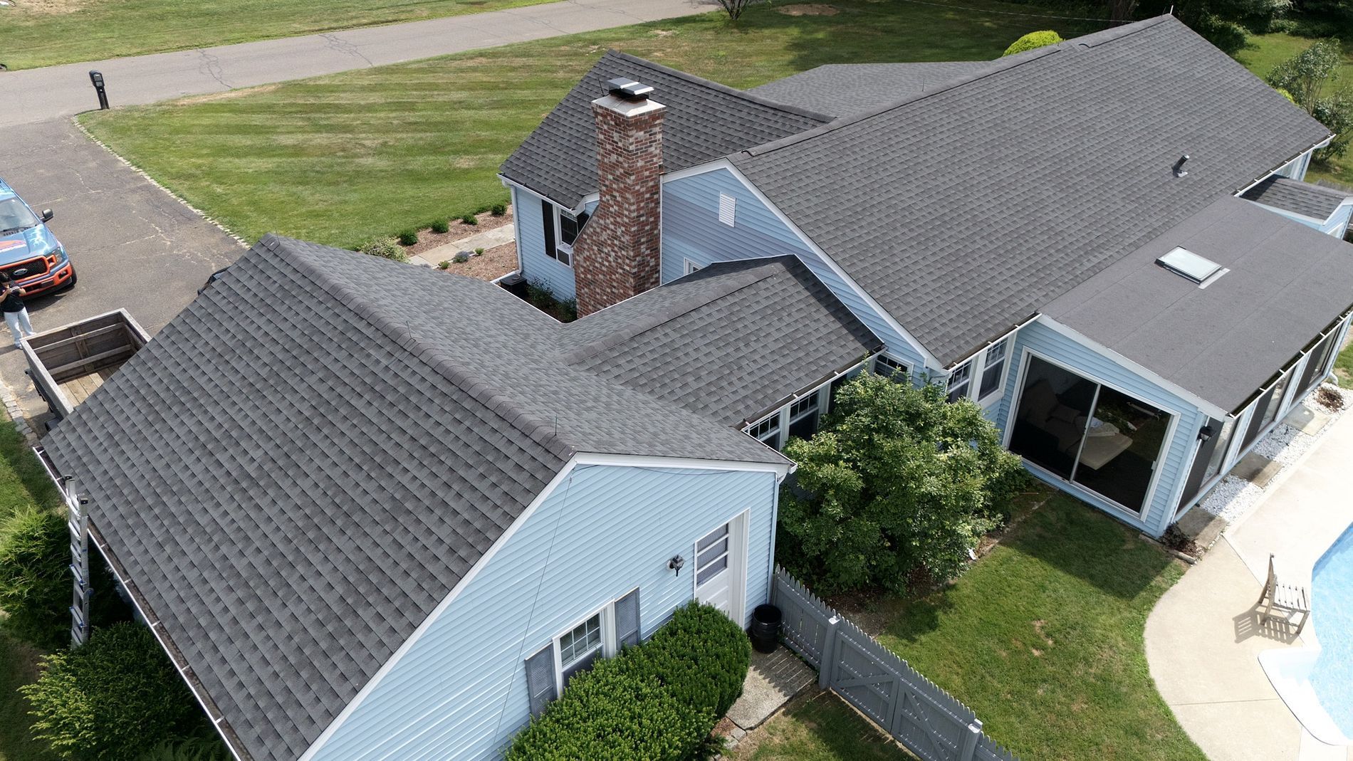 Aerial view of a suburban house with gray roofs, a brick chimney, and a landscaped yard
