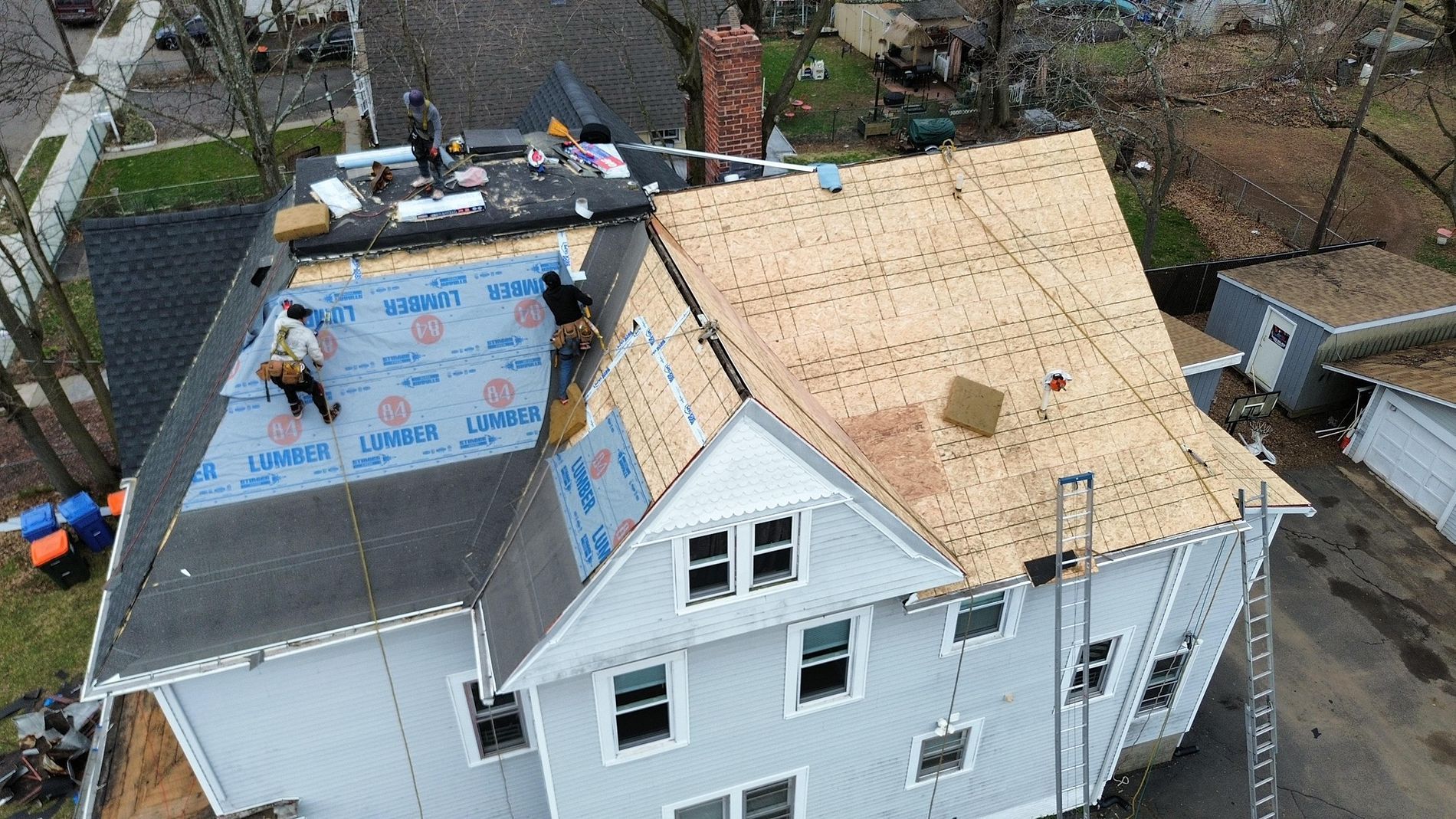 Aerial view of roof replacement on a white house, with workers on dark and plywood-covered sections.