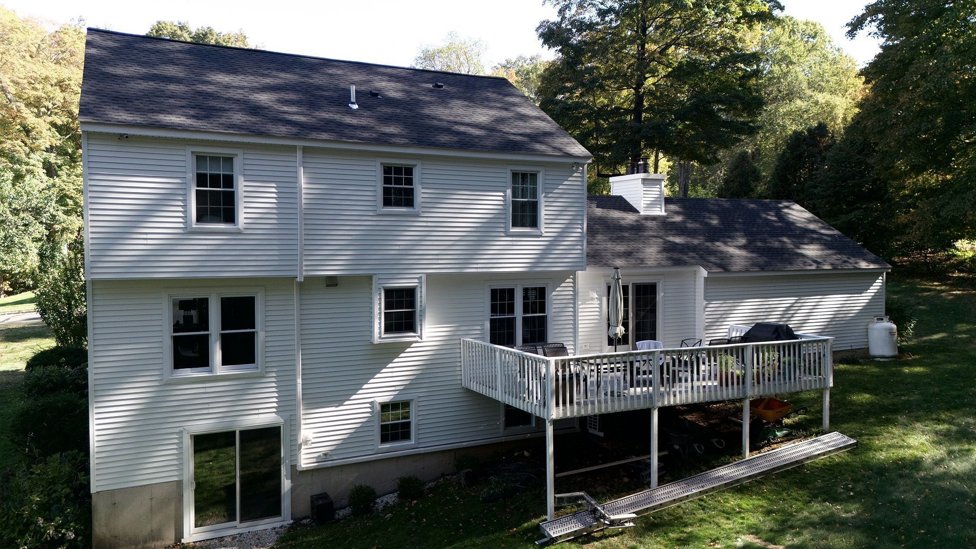 Two-story white house with a deck and sloped roof, surrounded by trees and lawn