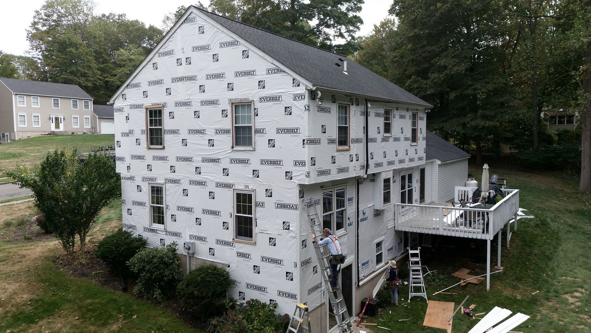 Partially wrapped house under construction with white housewrap and a raised deck in a wooded yard