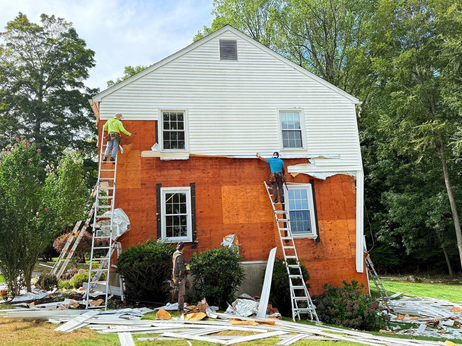 Workers on ladders repainting a red brick house with white siding and scattered paint supplies below
