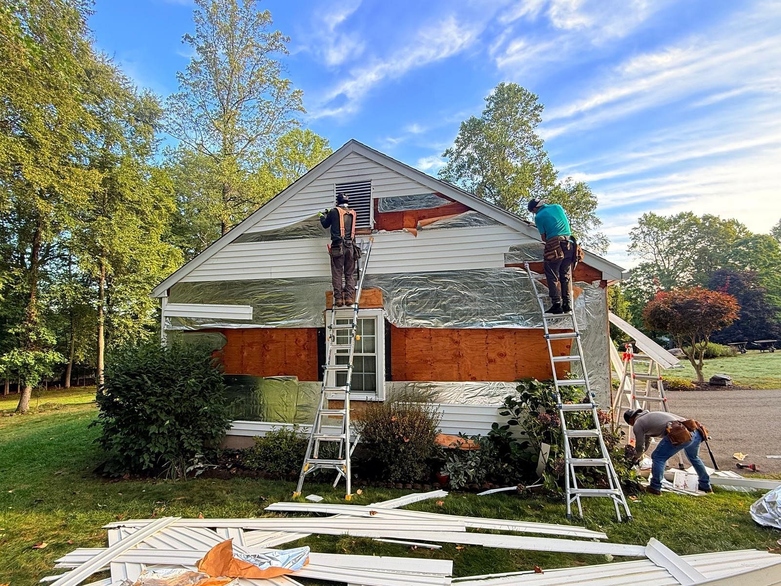 Workers on ladders painting a house exterior with white, red, and gray siding, with boards and tools on the lawn.
