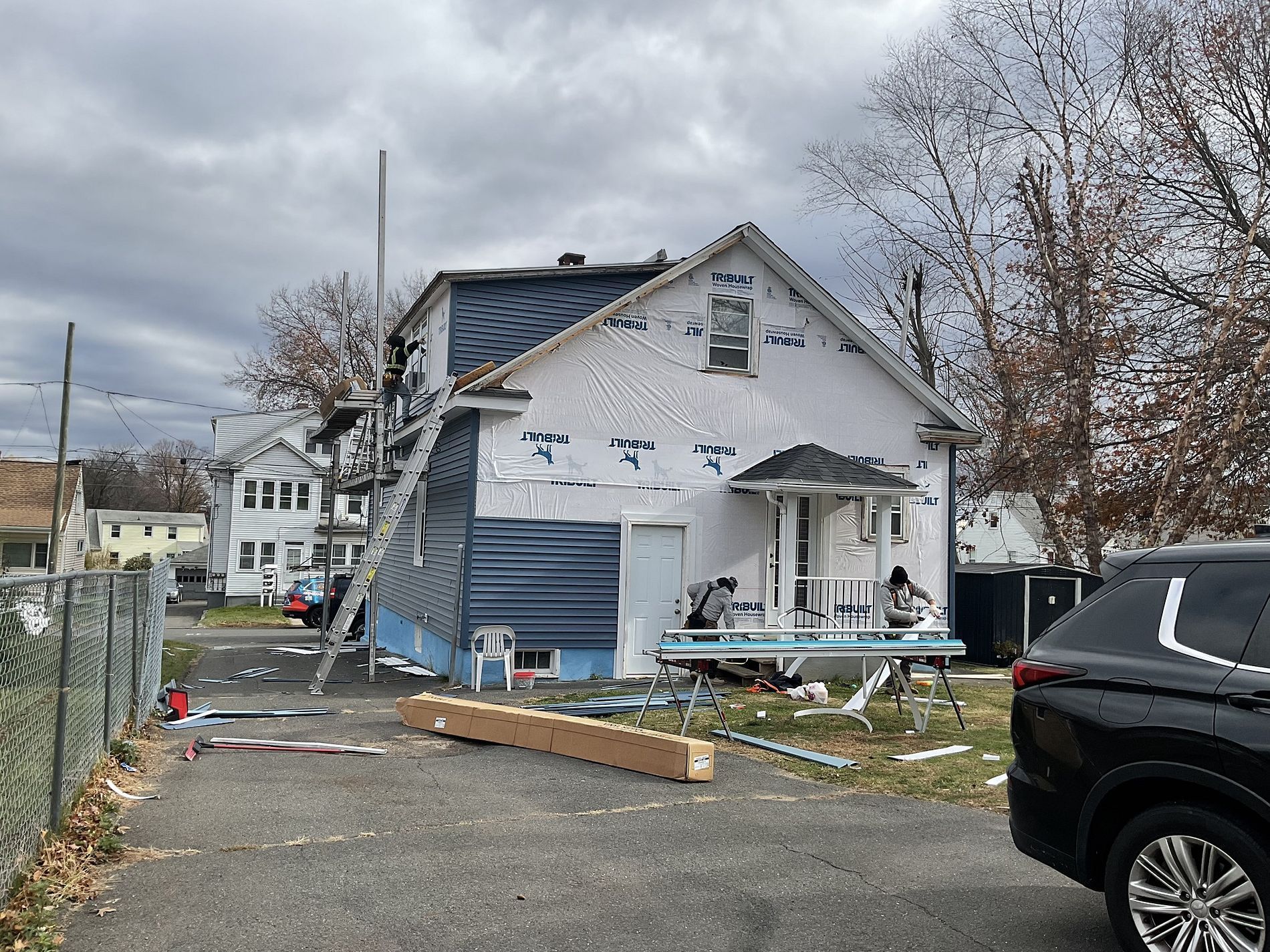 Damaged white house with debris in front and a black SUV parked at the curb