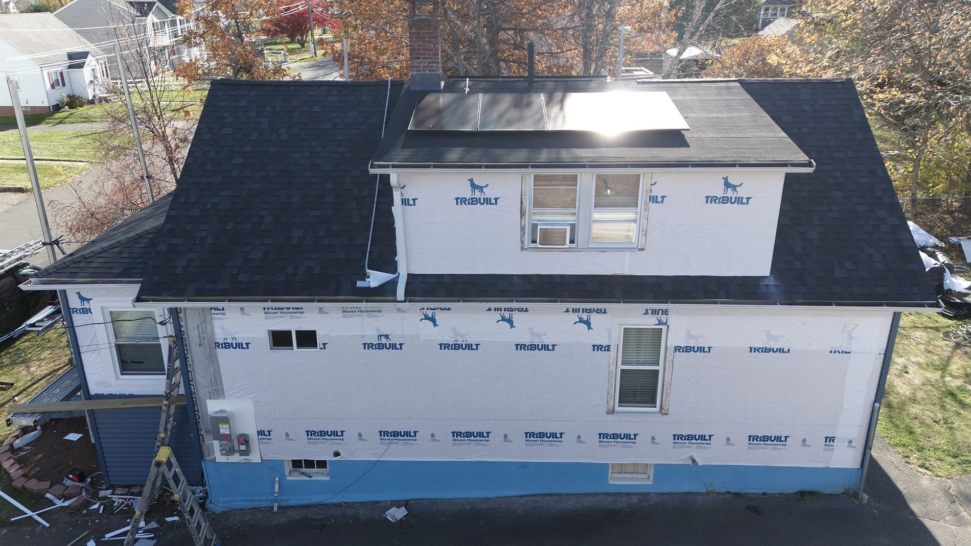A house under construction with white siding, black roof, and rooftop skylight in a suburban neighborhood