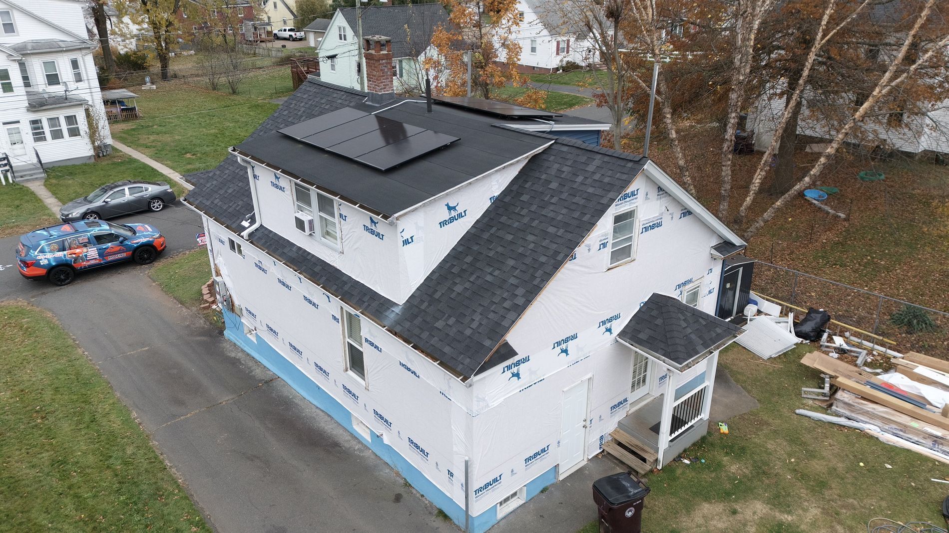 Aerial view of a white house with a dark roof, blue-trimmed lower level, and cars parked beside it.