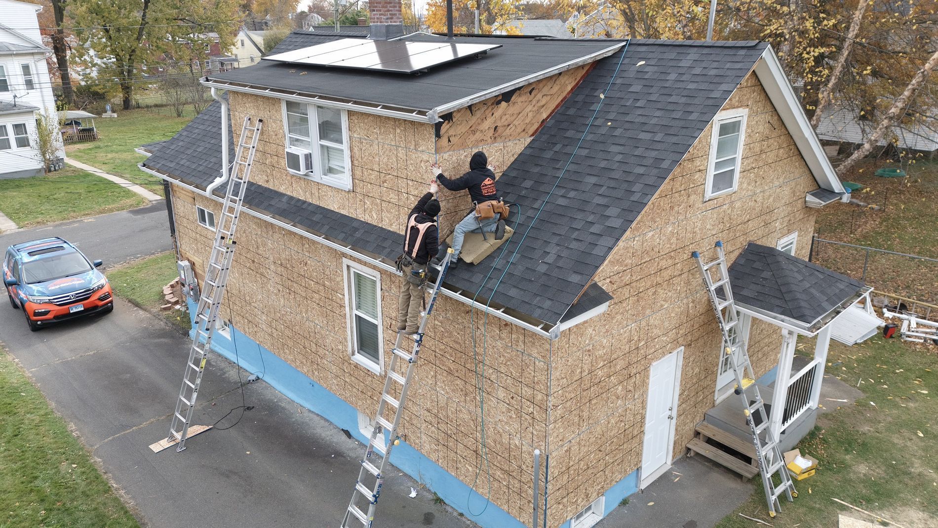 Worker on a steep roof repairing shingles on a brick house, with extension ladders below.