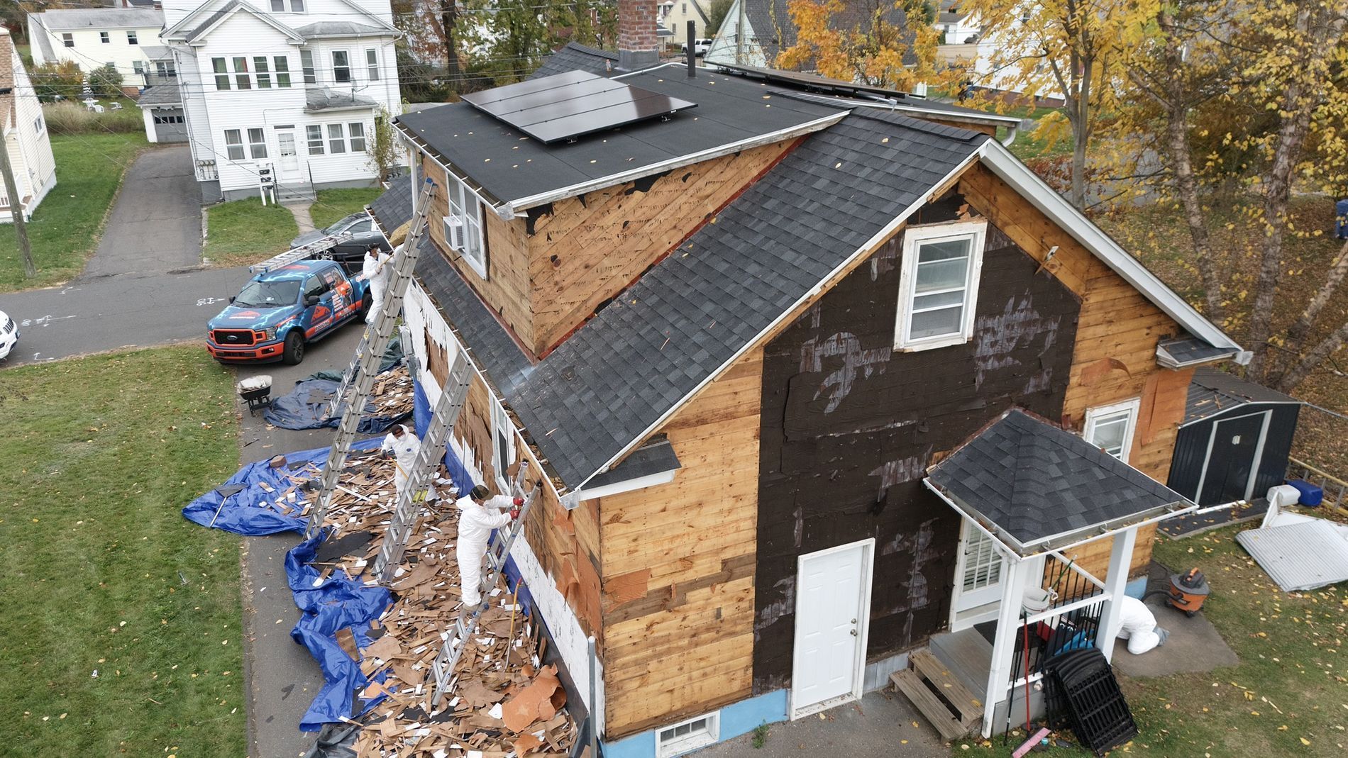 Aerial view of a damaged house with exposed roof, debris along the side, and a small porch.