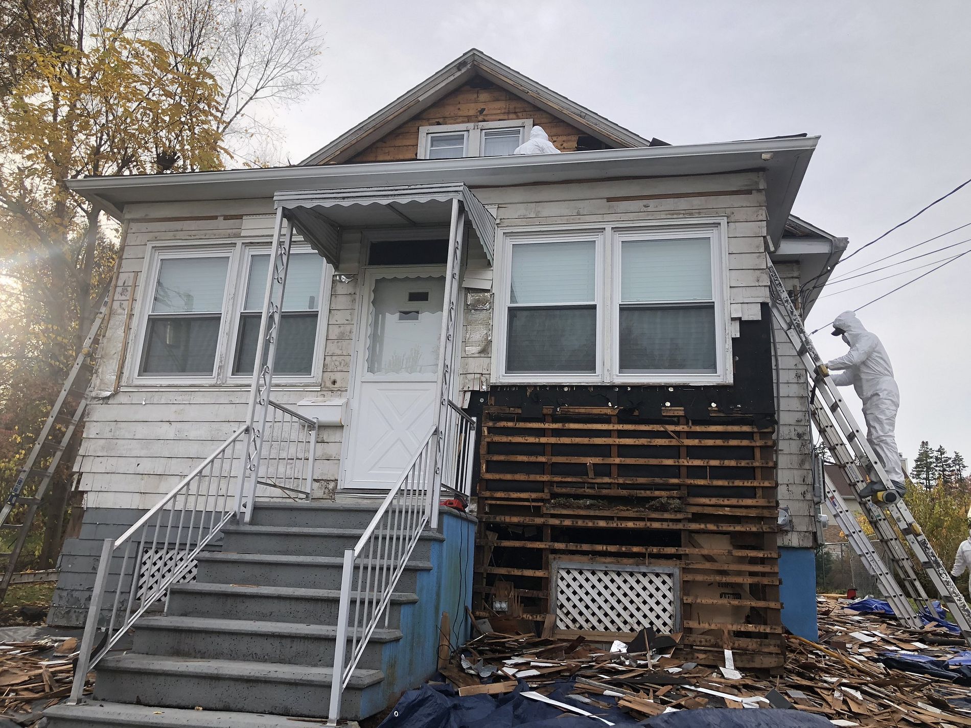 Gray house with front stairs and porch under repair, with exposed siding and debris below.