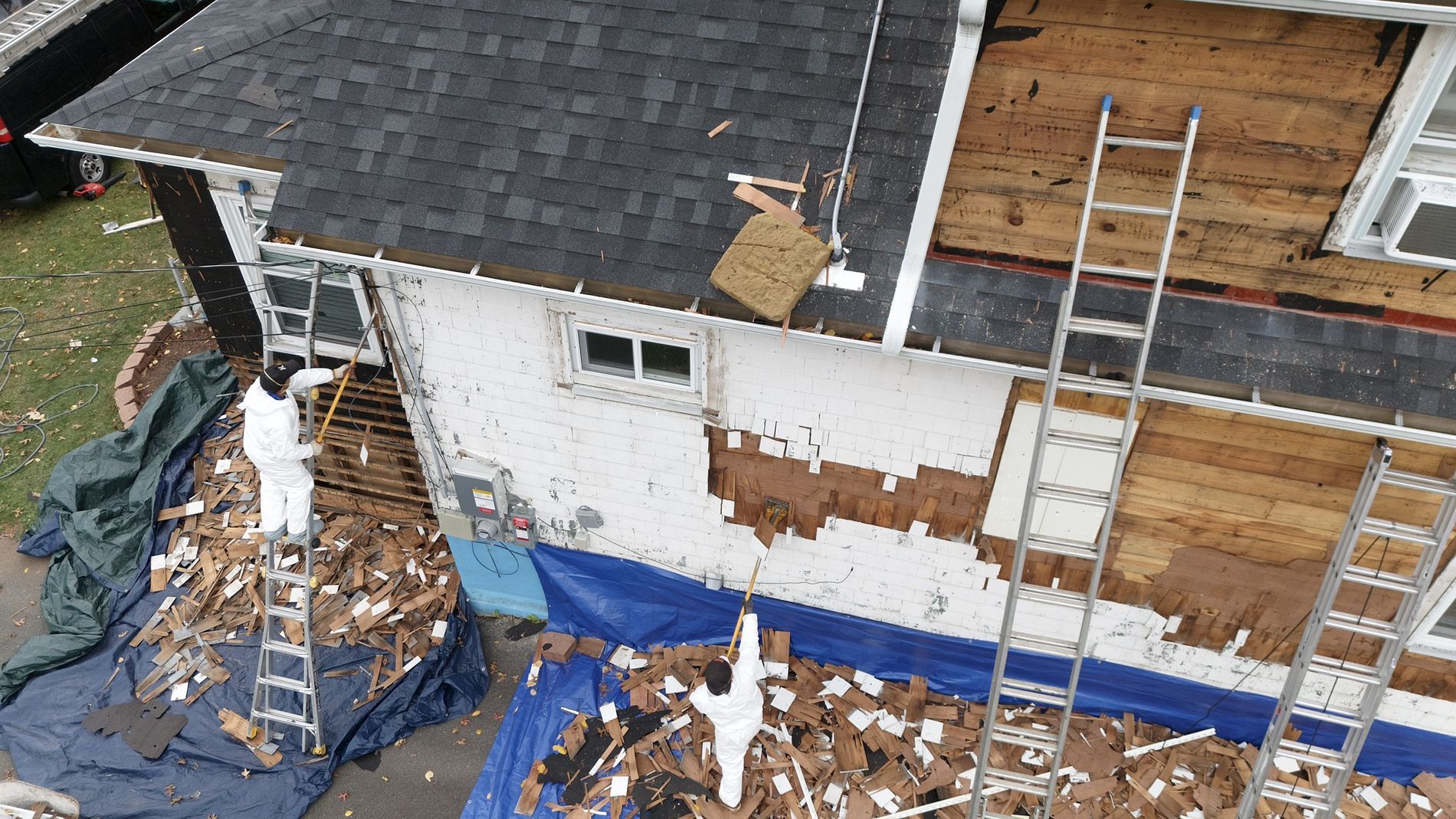 Roof repair on a house with exposed plywood, debris, ladders, and tarps surrounding the work area.