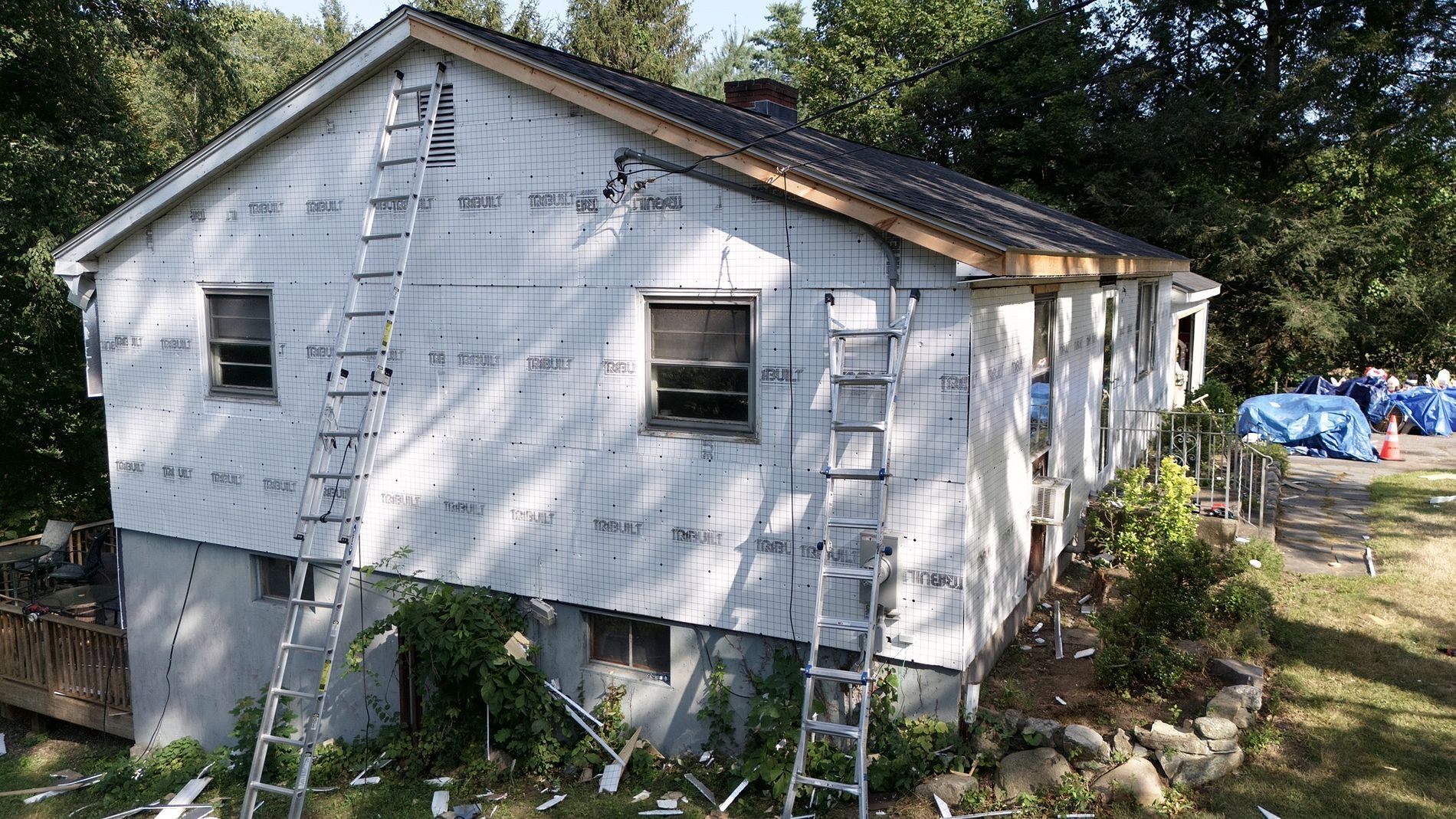 White house with ladder on the side, viewed from the yard with trees and construction materials nearby