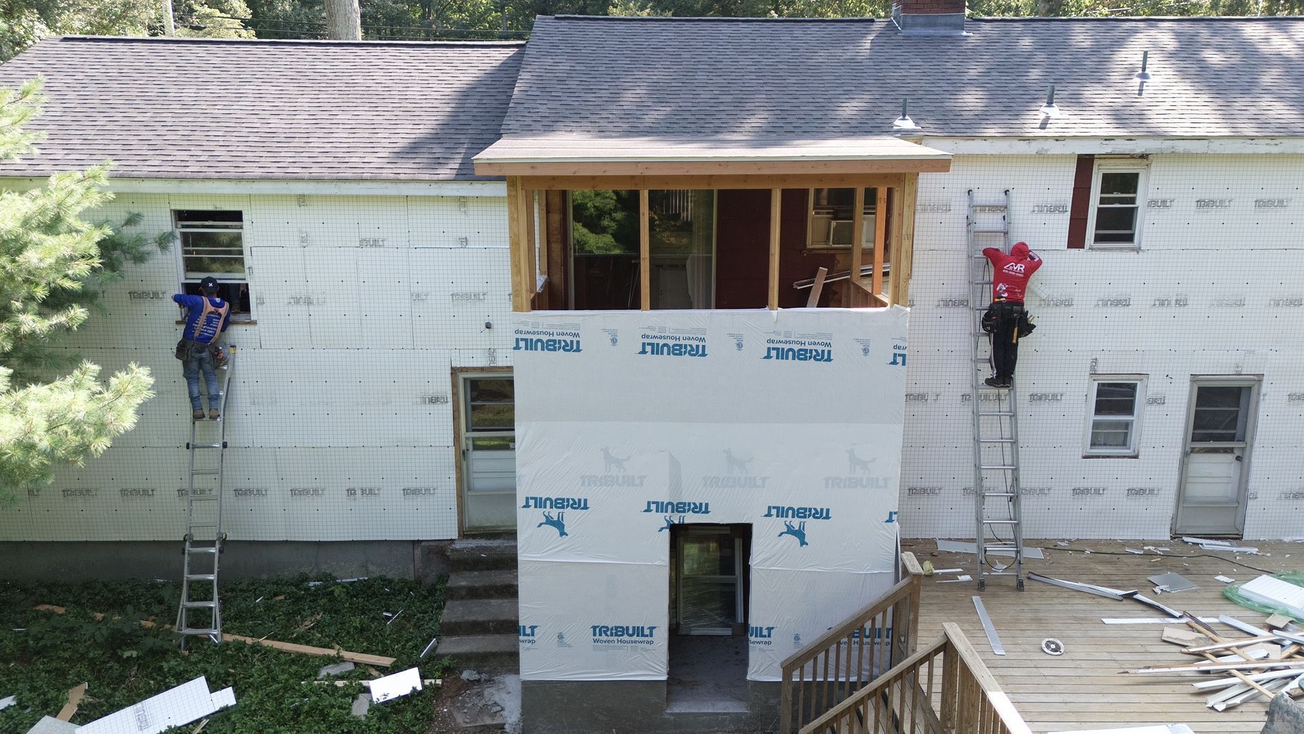 House exterior under renovation with workers on ladders, exposed insulation, and roofing in progress