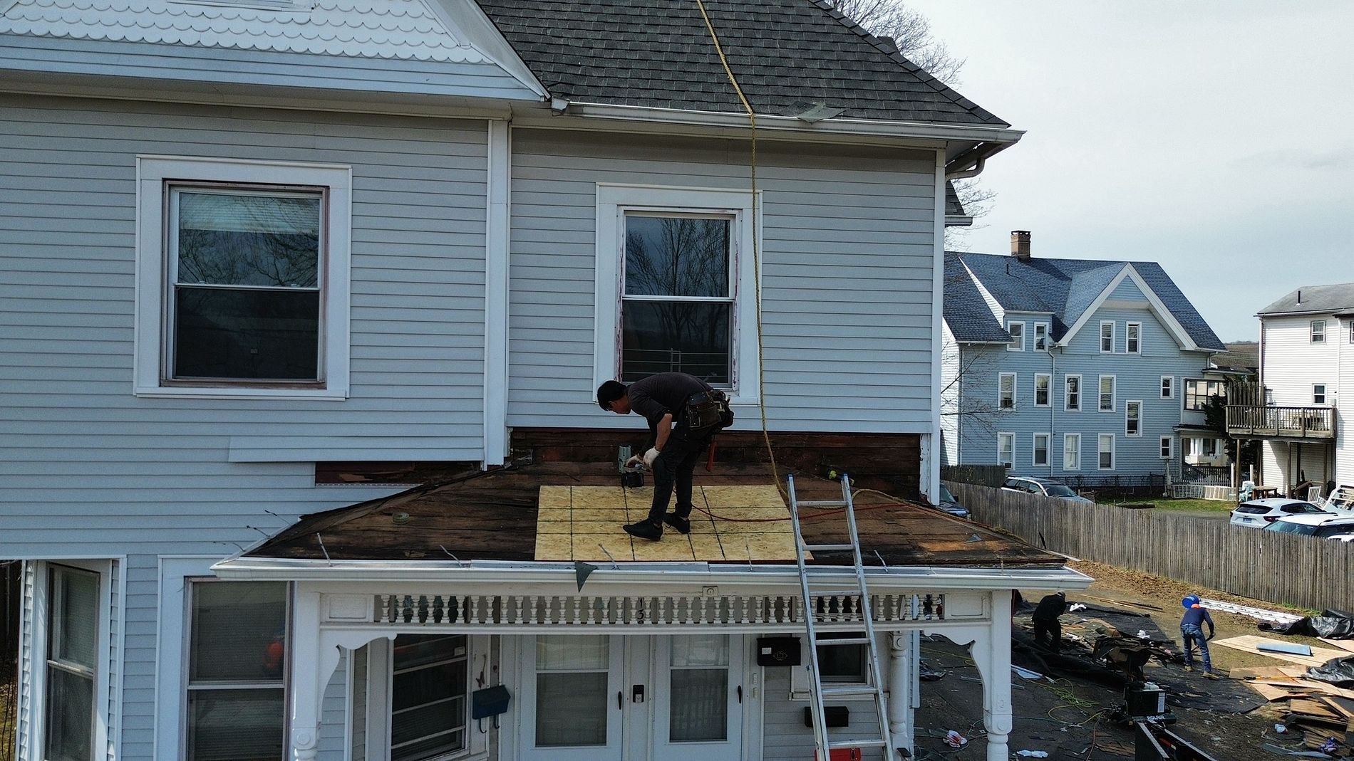 Worker on a house roof repairing shingles on a white building under daylight.