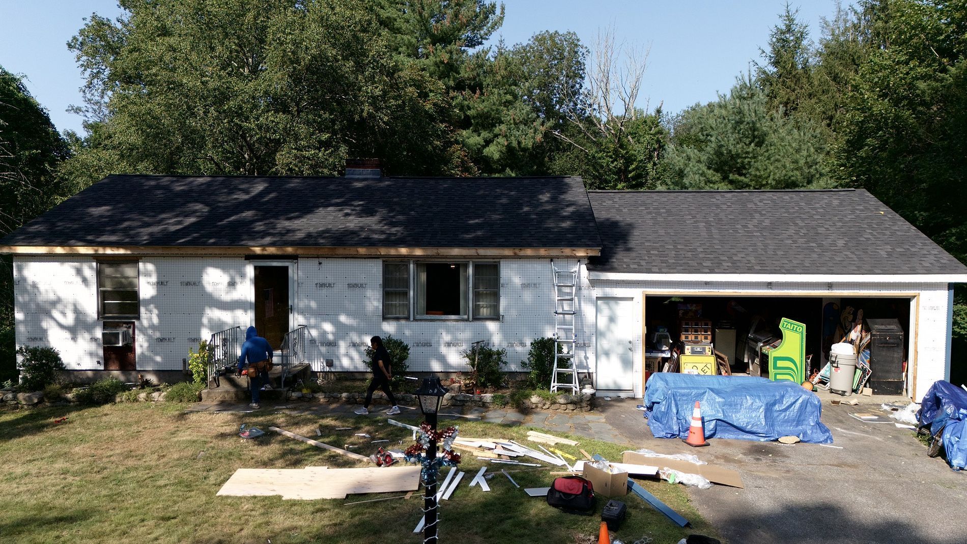 Single-story white house with a black roof and scattered yard equipment in the driveway.