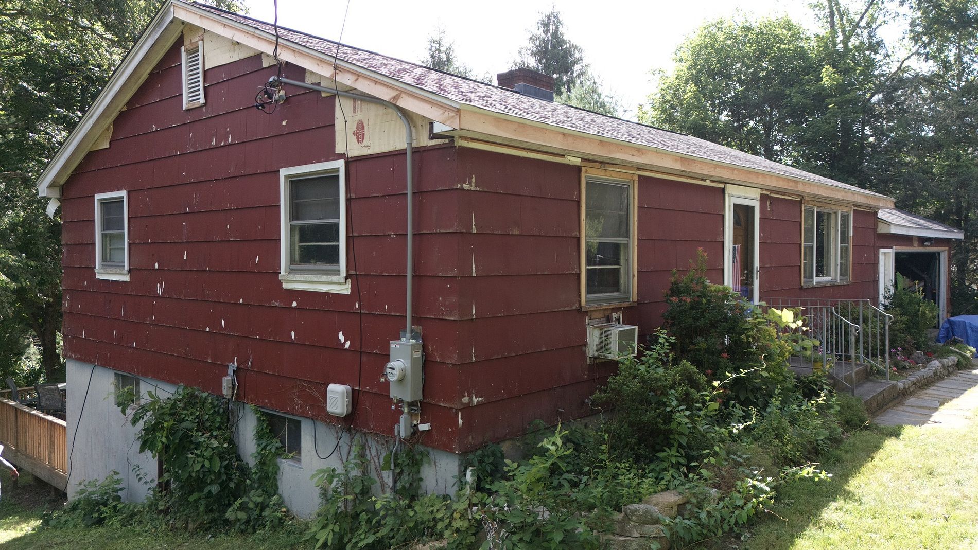Red house with white trim and a sloped roof, surrounded by overgrown greenery in a sunny yard.
