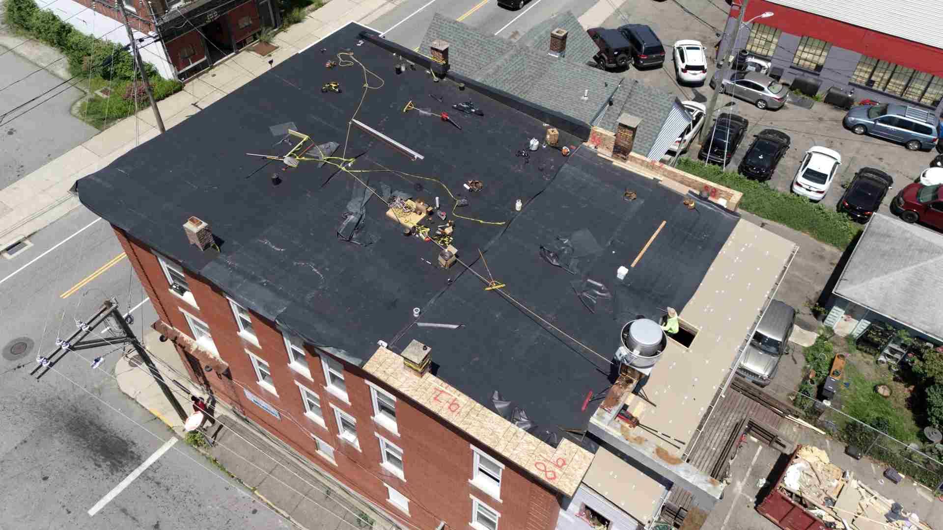 Aerial view of a flat black-roofed brick apartment building with HVAC units and nearby parked cars.