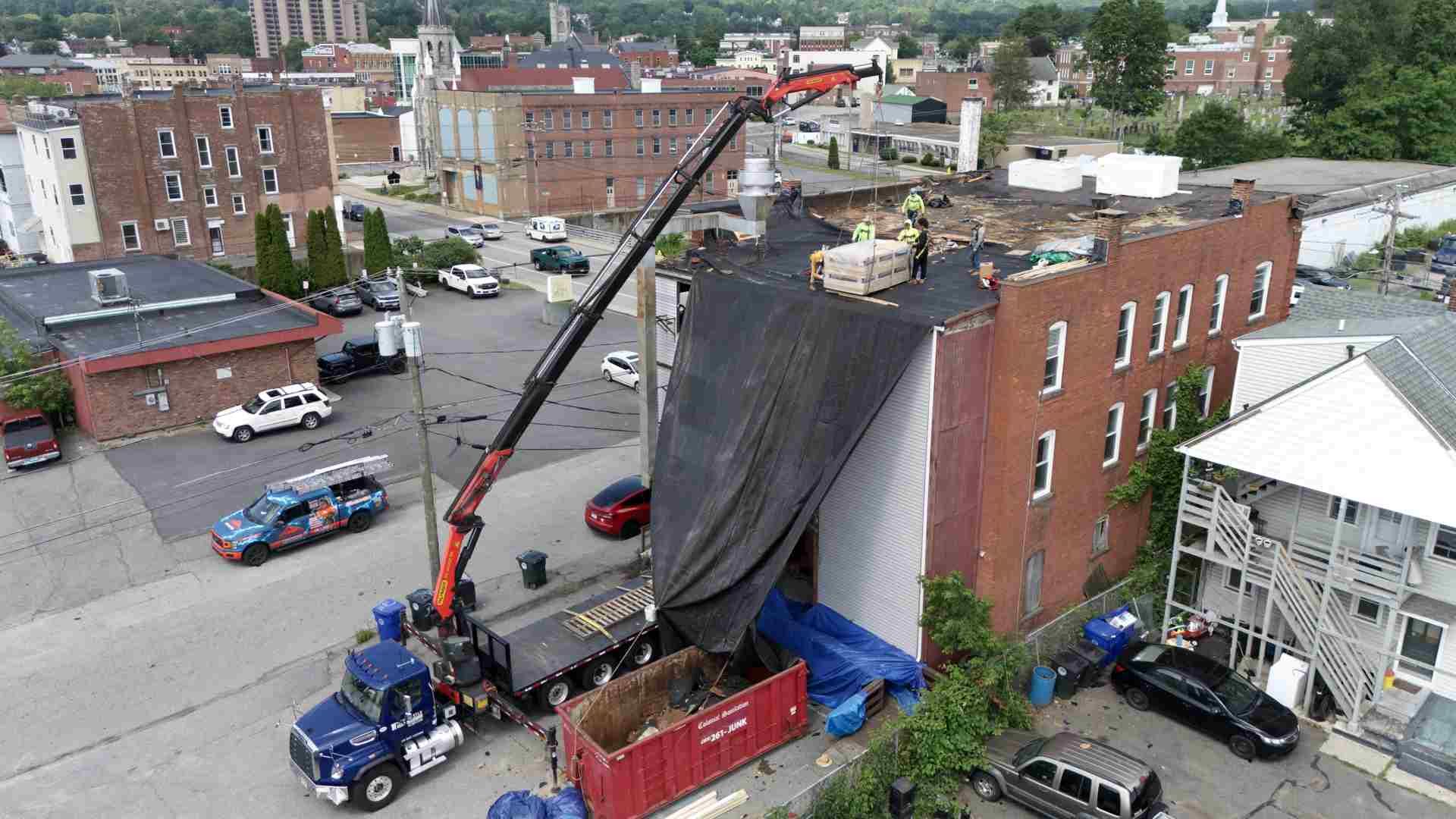 Crane demolishing a brick building on a city street beside parked cars and a red dumpster