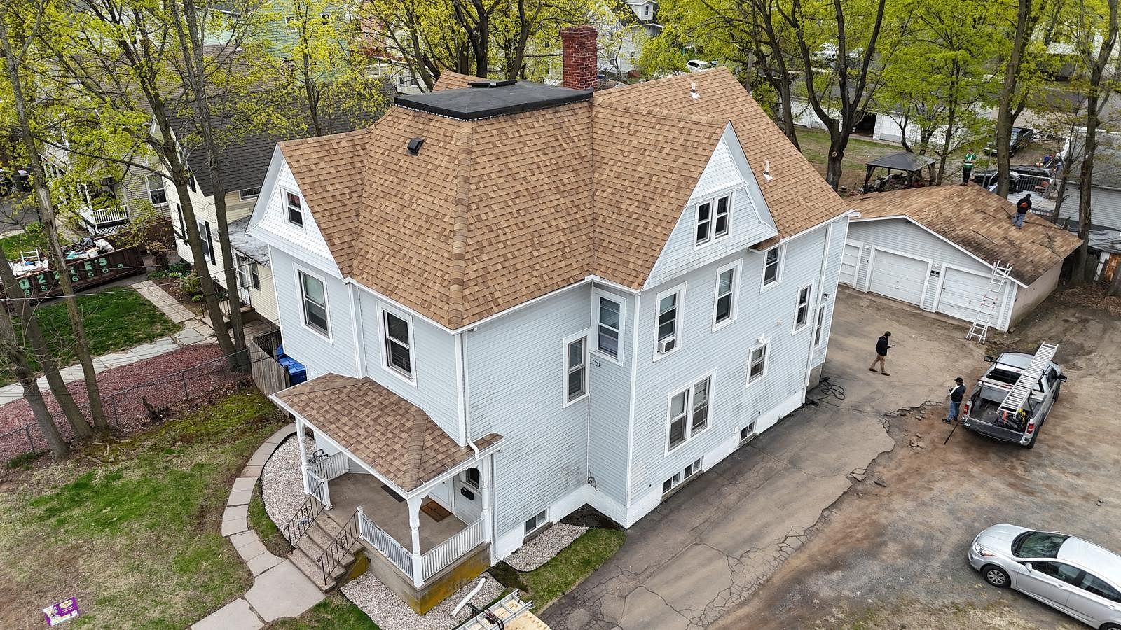 Aerial view of a white house with a brown roof, porch, detached garage, and cars in the driveway.