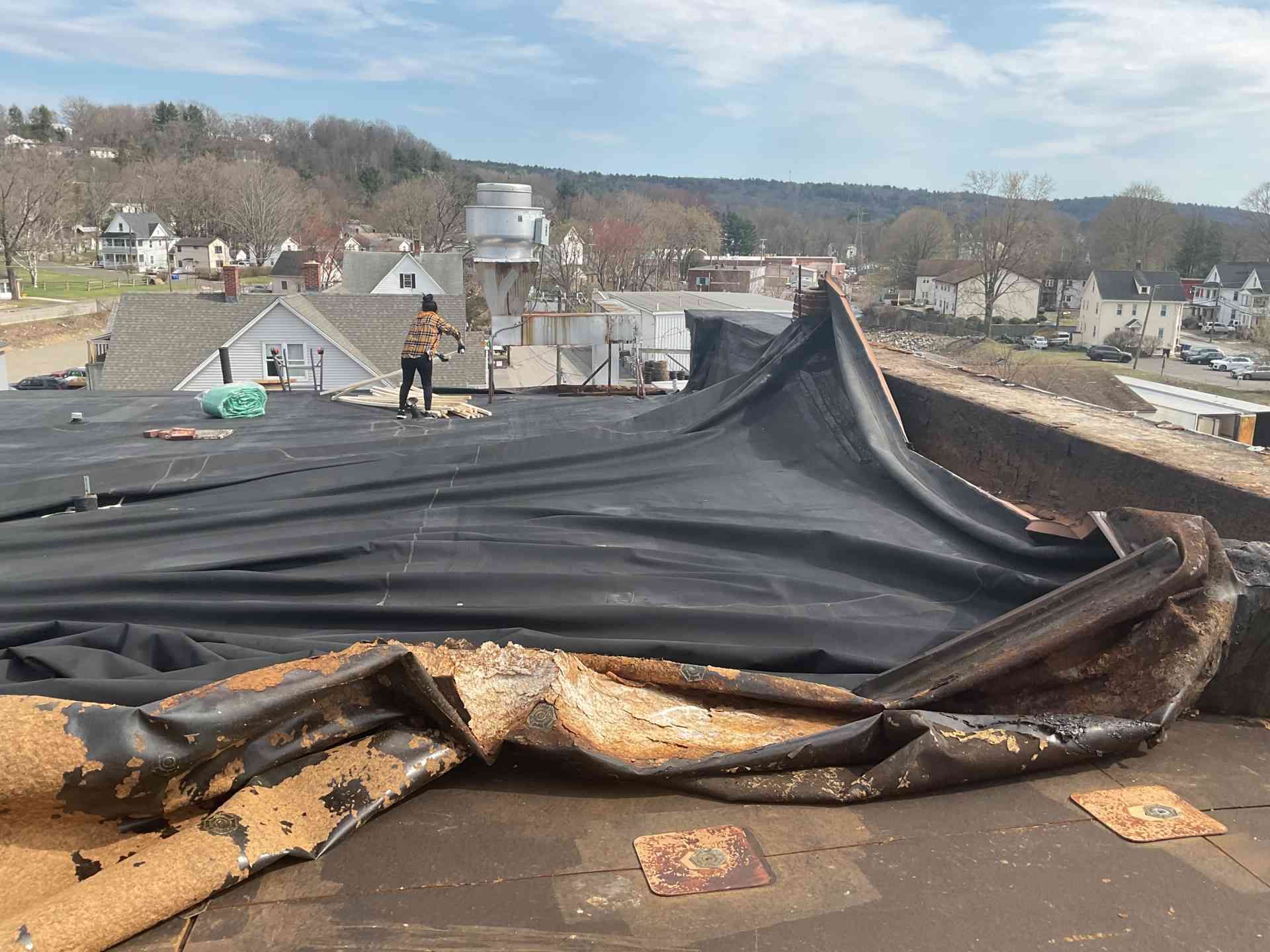 Ripped black roof membrane on a flat rooftop, with a person working near damaged edges and a neighborhood in the background