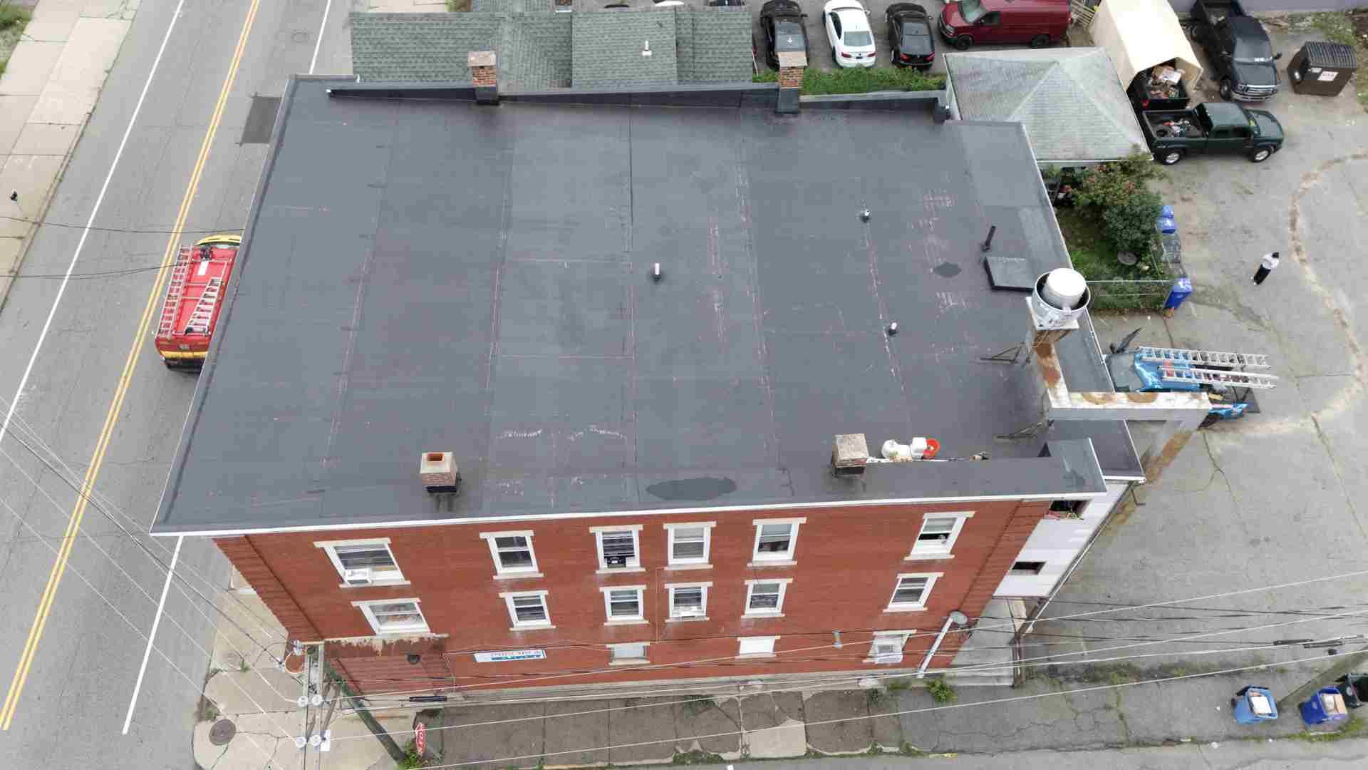 Aerial view of a red brick building with a flat dark roof beside a street and parking lot.