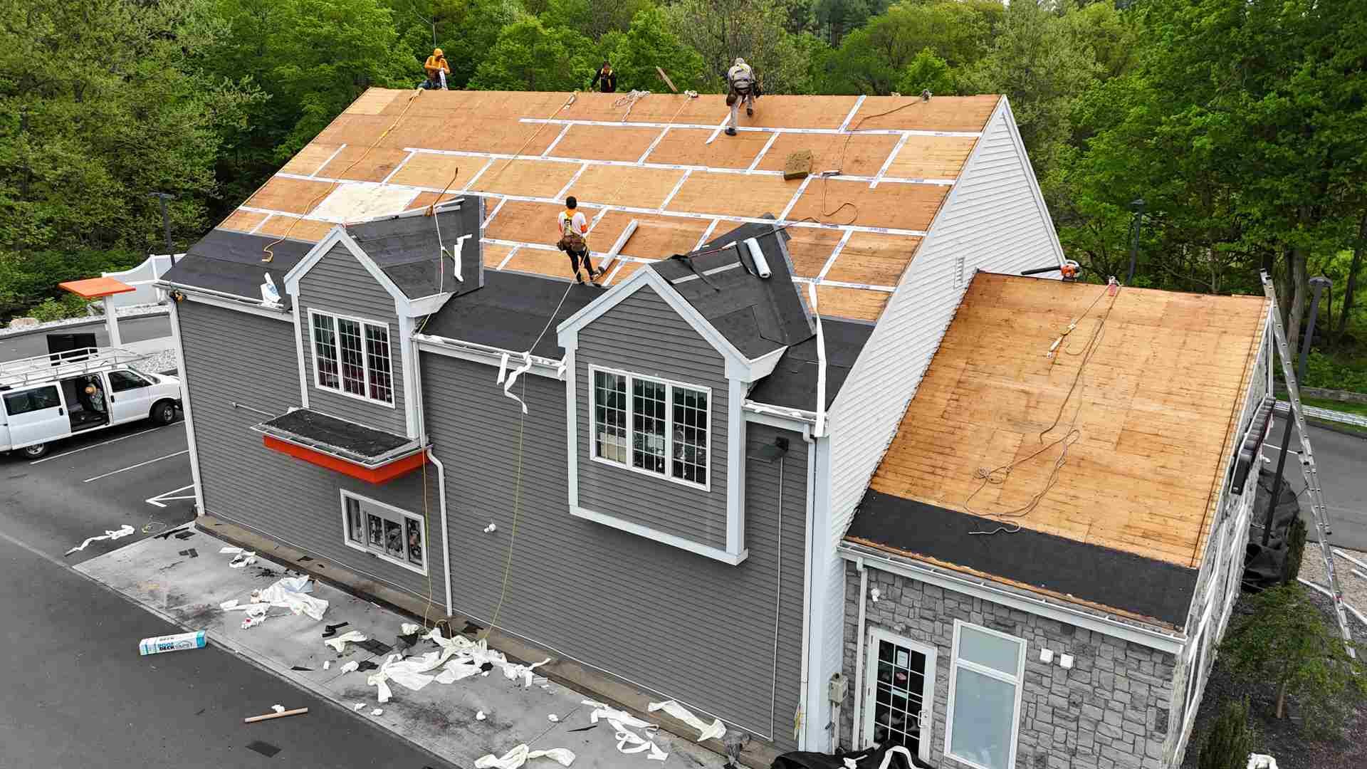 Aerial view of roofers working on a gray house roof with exposed plywood and partially replaced shingles.