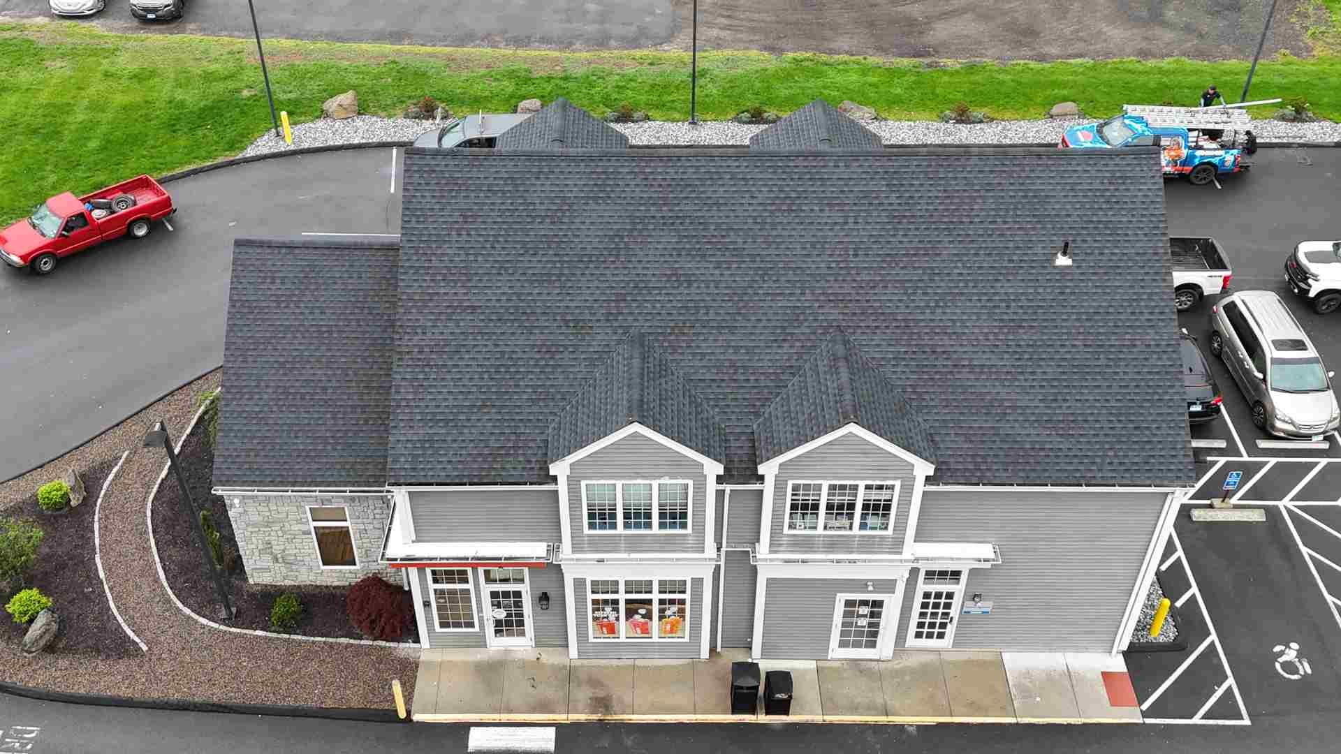 Aerial view of a gray storefront building with dark roof, parking lot, and a few cars nearby