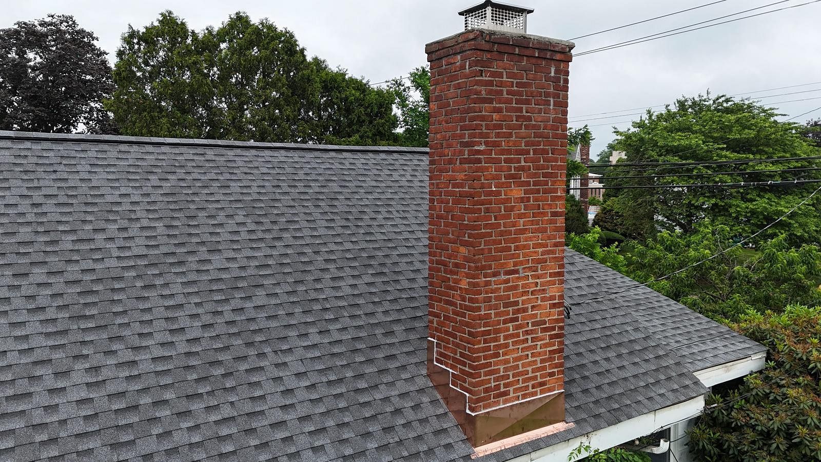 Gray shingled roof with a red brick chimney, surrounded by trees