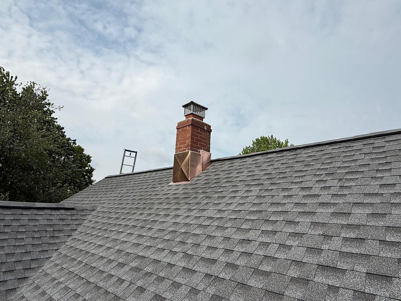 Gray shingled roof with a brick chimney under a cloudy sky, trees at the left edge