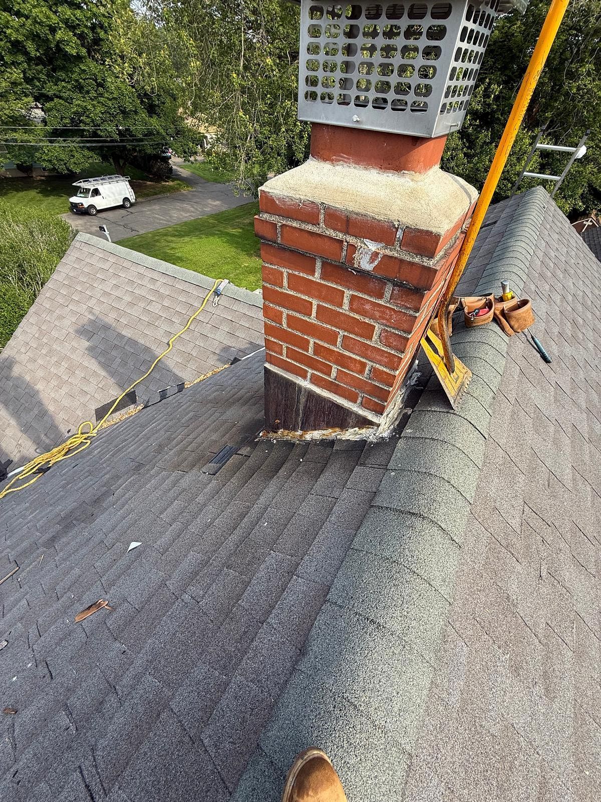 Rooftop chimney with red brick base and white vent cap, beside gray shingles and a yellow safety rope.