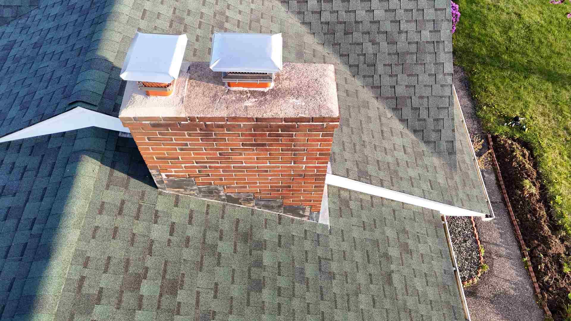 Rooftop view of a brick chimney with two metal caps on green shingles.
